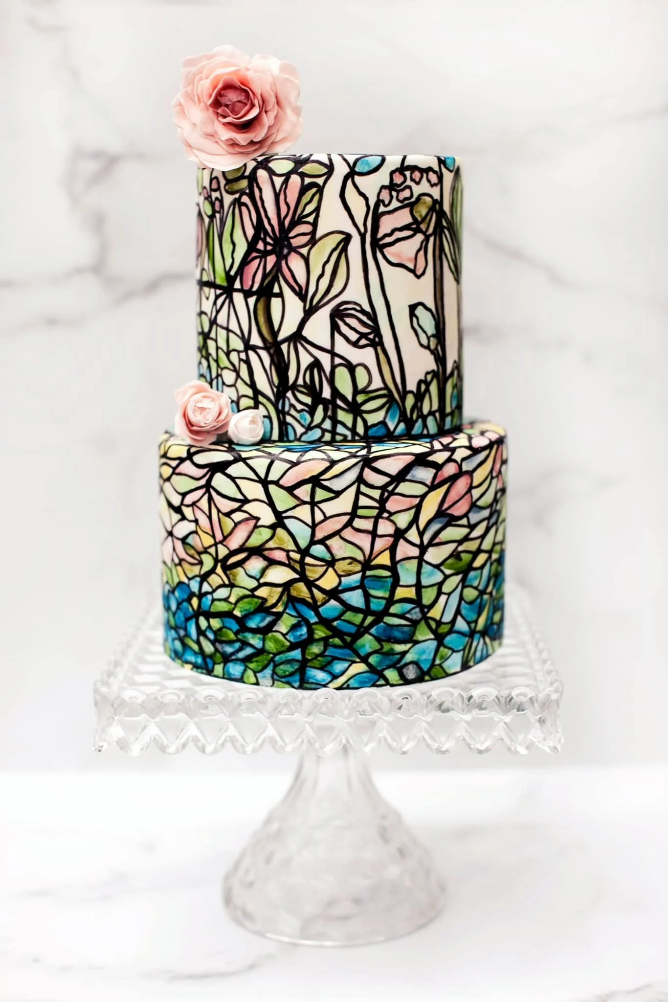 Two-tiered cake decorated with a stained glass floral pattern and pink roses on top, displayed on a glass cake stand.