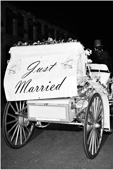 A vintage horse-drawn carriage with a large sign that says "Just Married" and floral decorations, seen at night with a person wearing a top hat riding it.