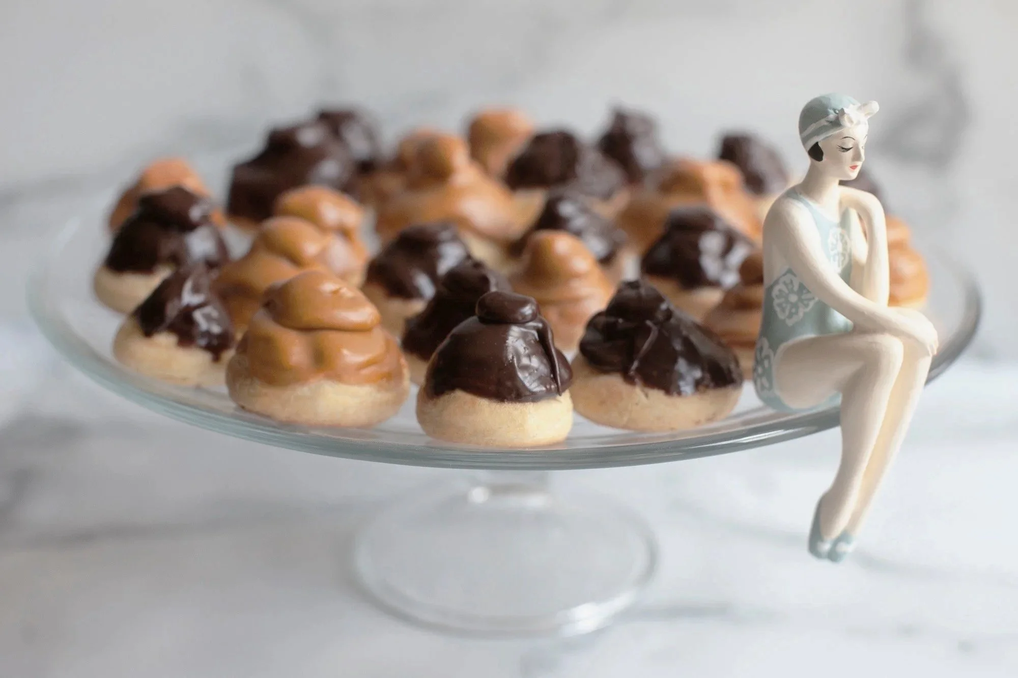 Assorted cookies with chocolate and caramel toppings on a glass serving tray, with a decorative figurine of a woman sitting on the side of the tray.