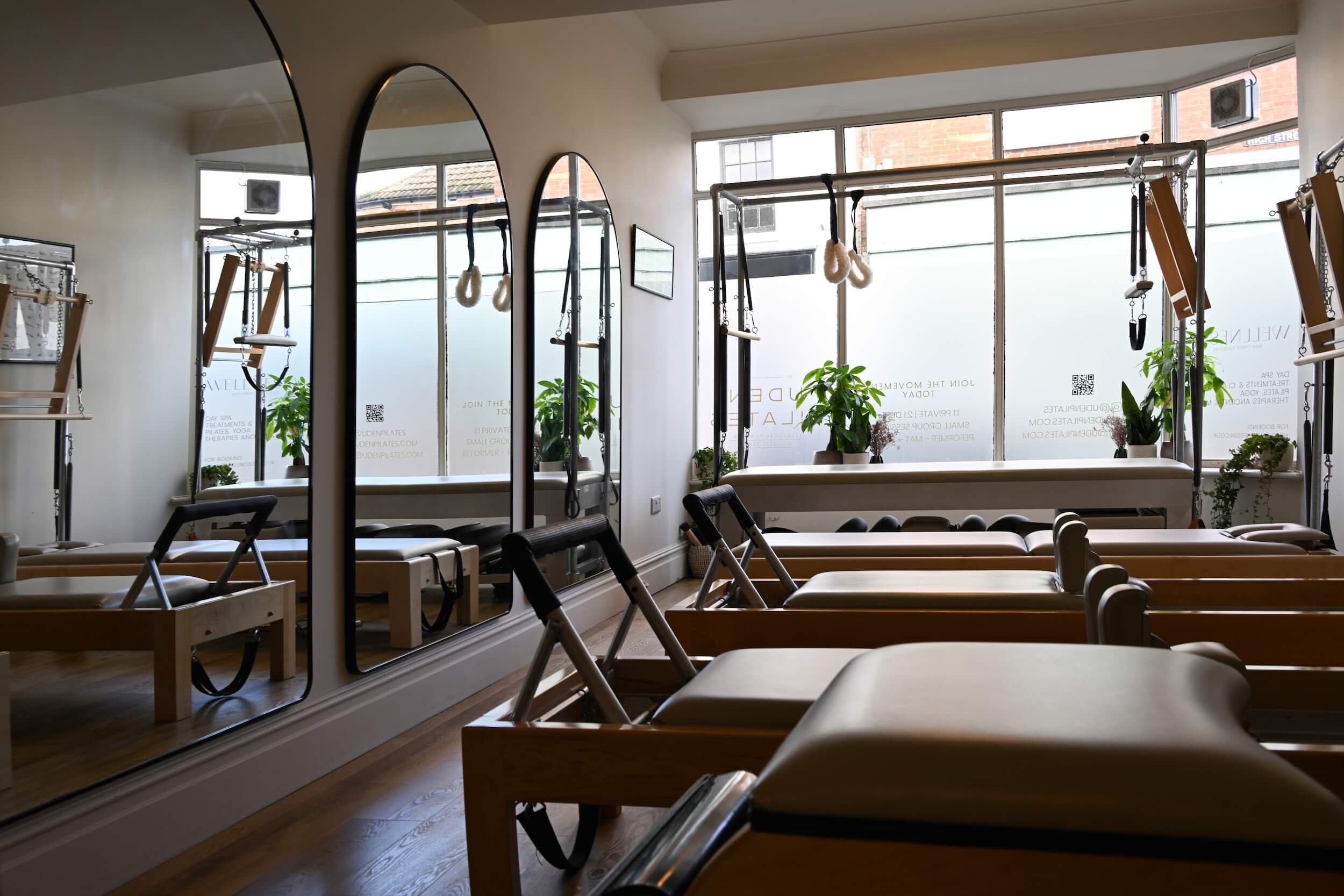 A Pilates or fitness studio with wooden reformer machines, gymnastic rings, resistance bands, and mirrors, illuminated by natural light from large windows decorated with potted plants.