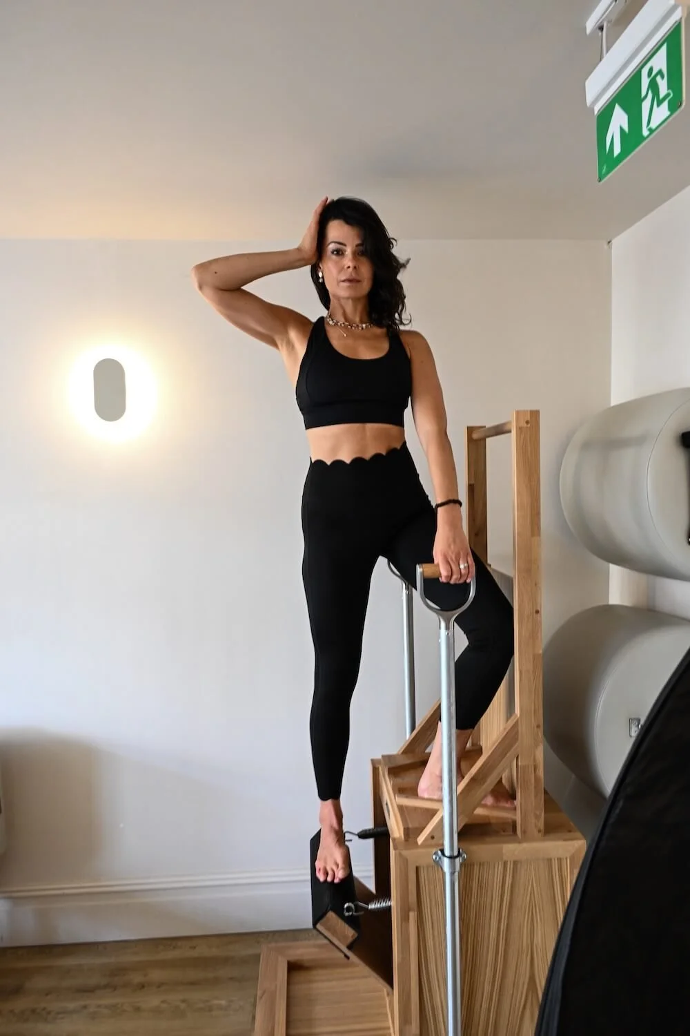 A woman dressed in black workout attire standing on a Reformer pilates device, with one foot on the device and the other foot on a pedal, inside a room with white walls and a white ceiling.