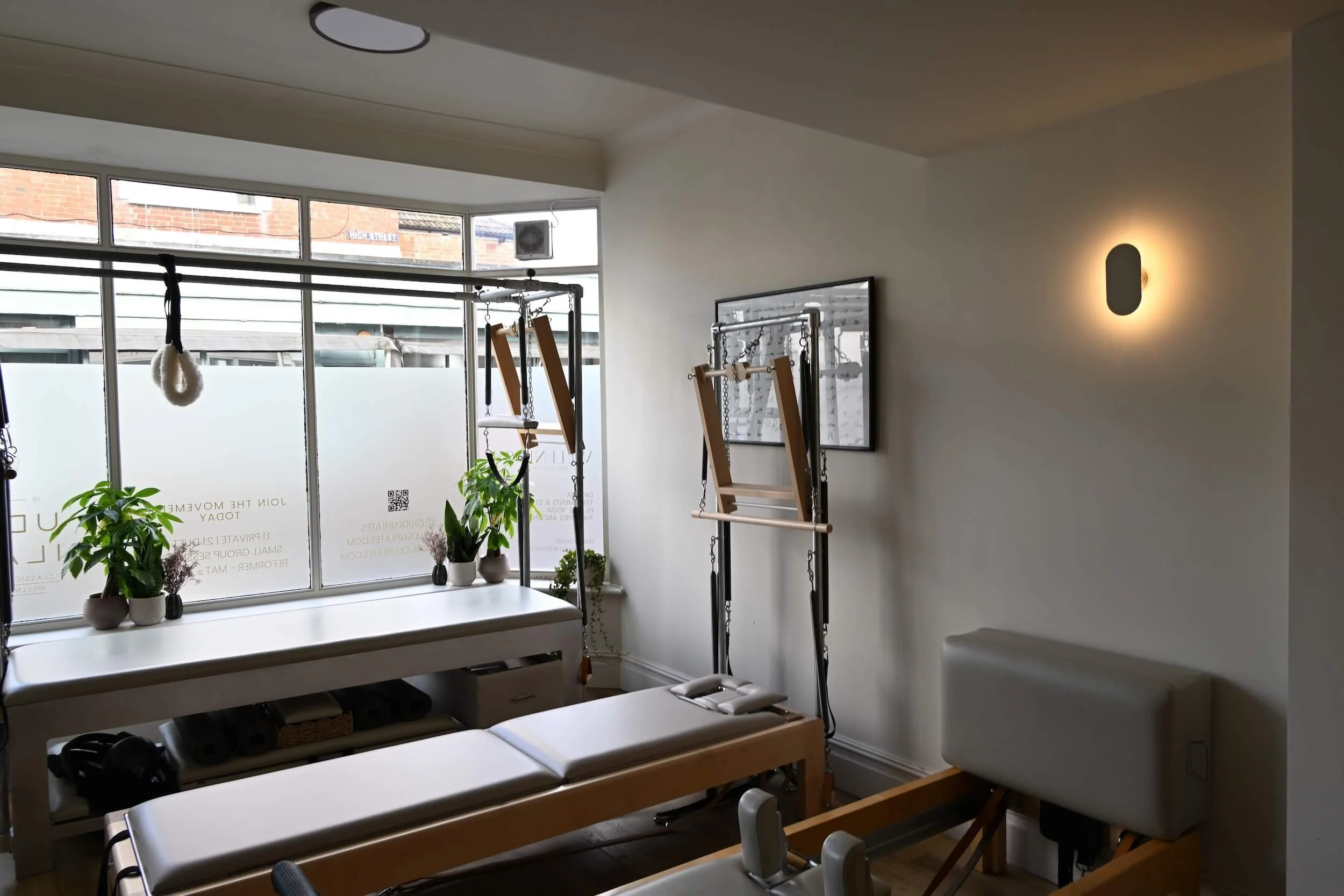Interior of Uden Pilates fitness studio featuring Pilates equipment, a large window with plants, wall-mounted light, and exercise frames.