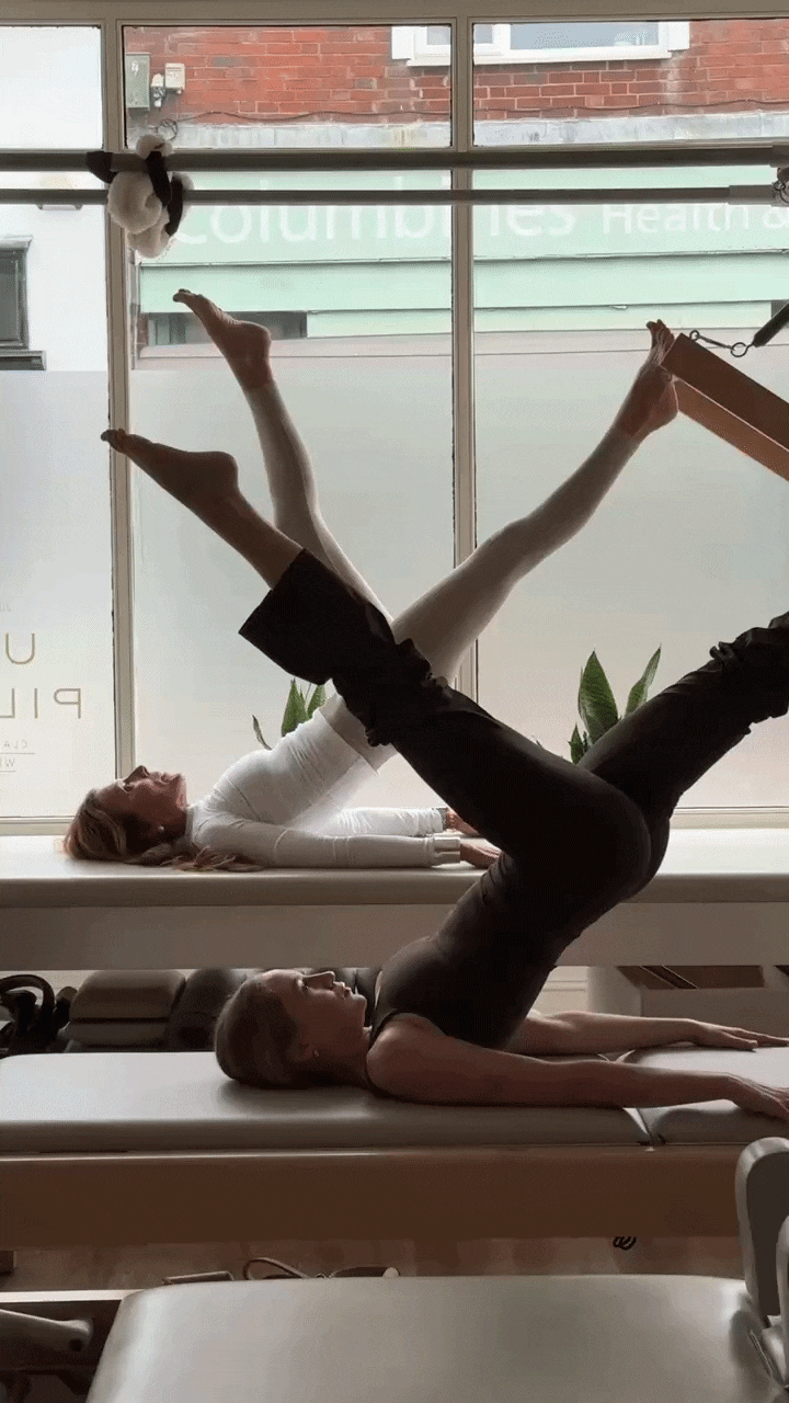 2:1 Duet Session, two women practicing pilates on a windowsill, with one lying on her back and lifting her legs and arms, while the other maintains a similar pose. Sunlight filters through large windows behind them.