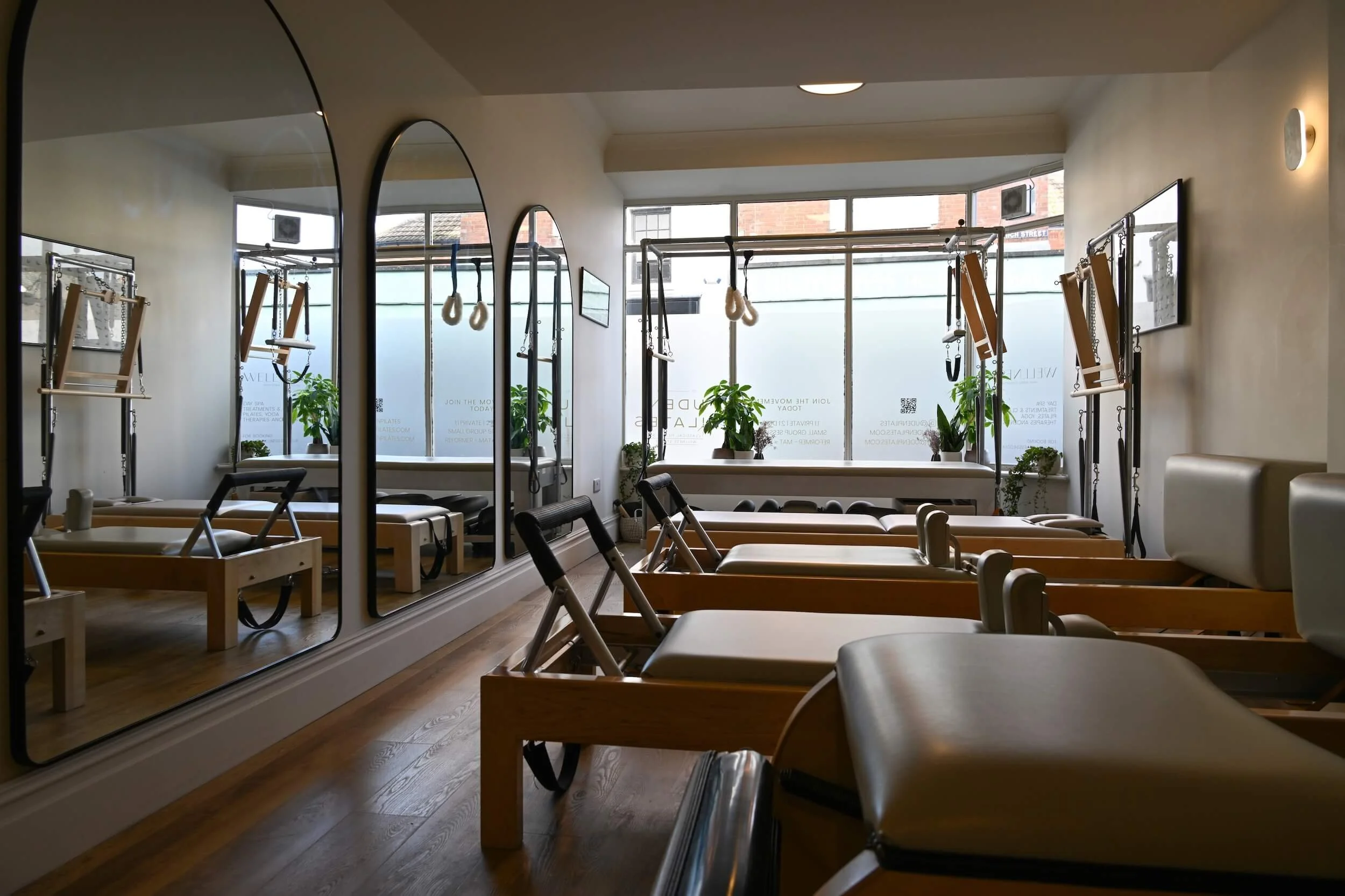 Uden Pilates studio with reformer machines, rings, and mirrors, illuminated by natural light from large front windows with potted plants.