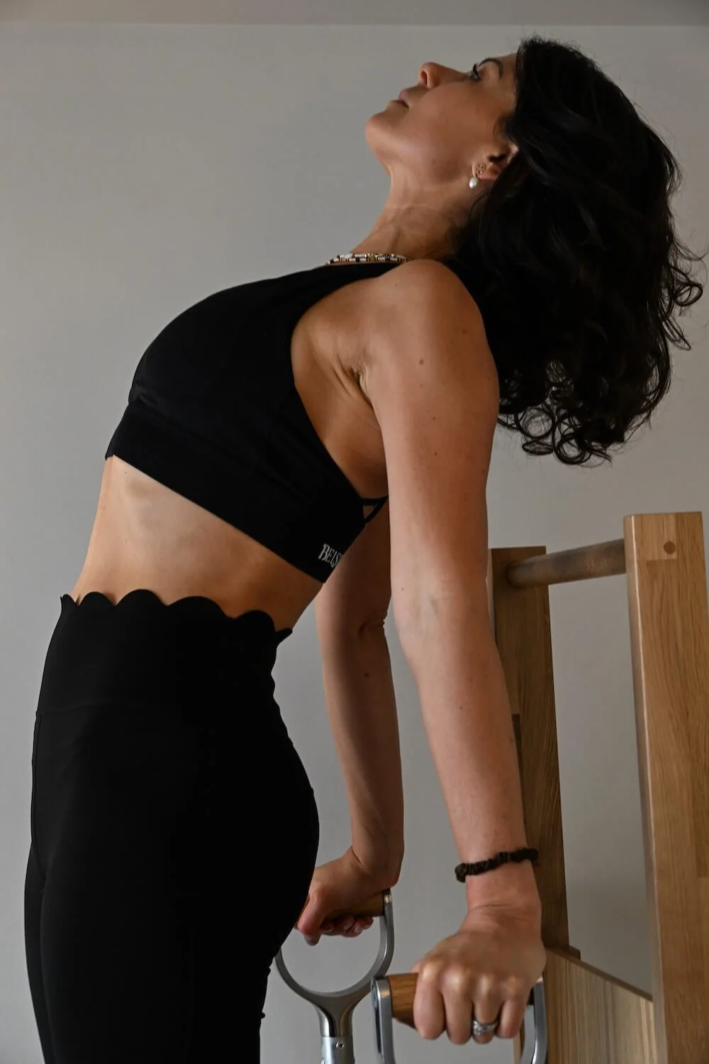 A woman doing a fitness exercise using parallel bars, wearing a black sports top and black pants, with dark wavy hair and earrings.