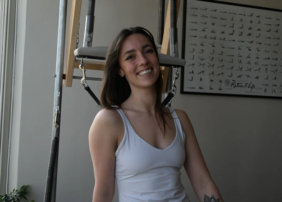 Woman smiling in front of a wall chart with illustrations of bird poses, standing next to an indoor exercise equipment with a padded seat.