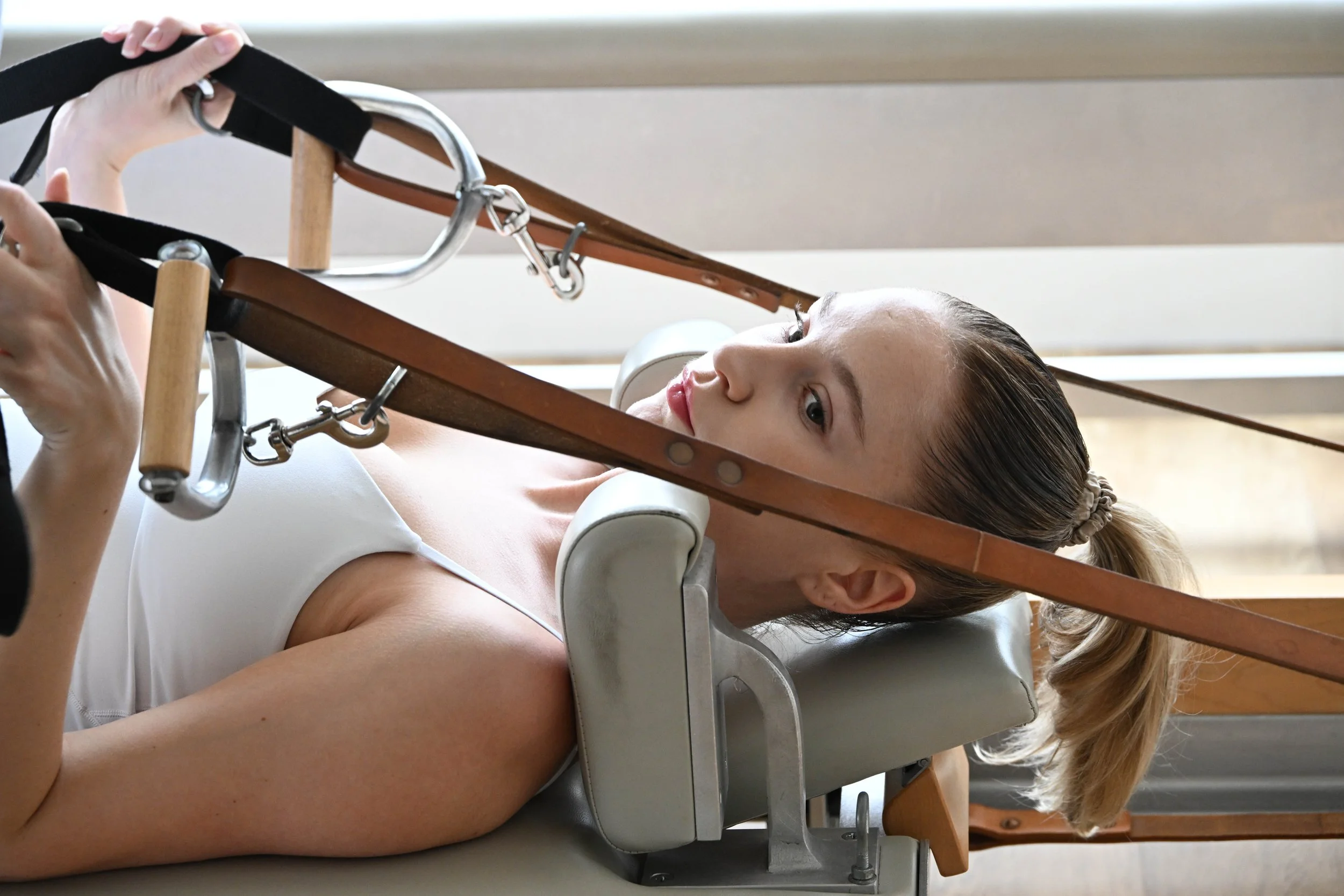 A woman doing Pilates using a reformer pilates machine, lying on her back with her head supported and straps holding her head in place.