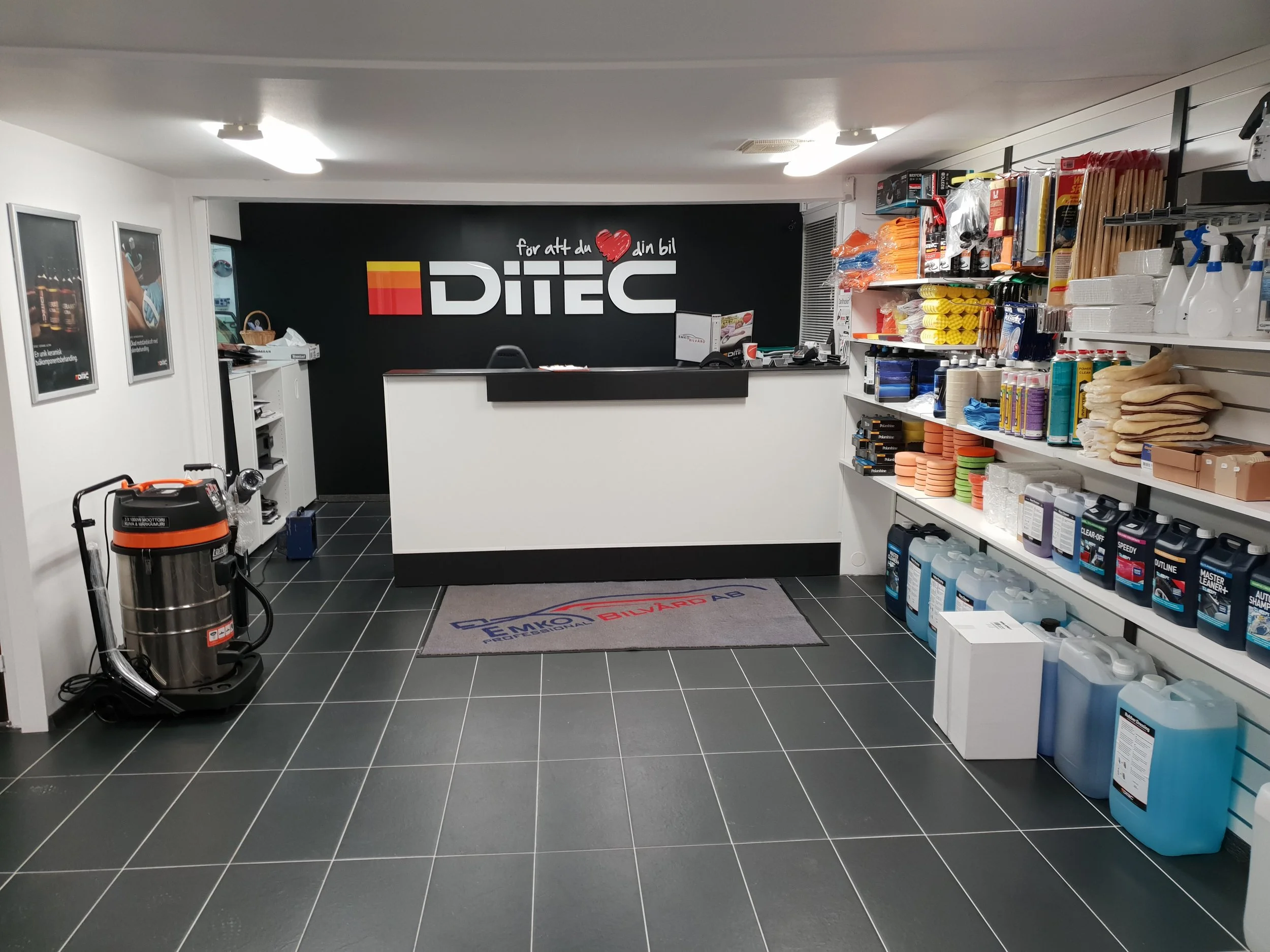 Inredning av en butik eller mottagning med en svart vägg bakom ett kassabord, hyllor fyllda med rengöringsprodukter, sopborstar och andra städhjälpmedel. En matta framför kassabordet med företagslogga och en industrivakuummaskin till vänster.