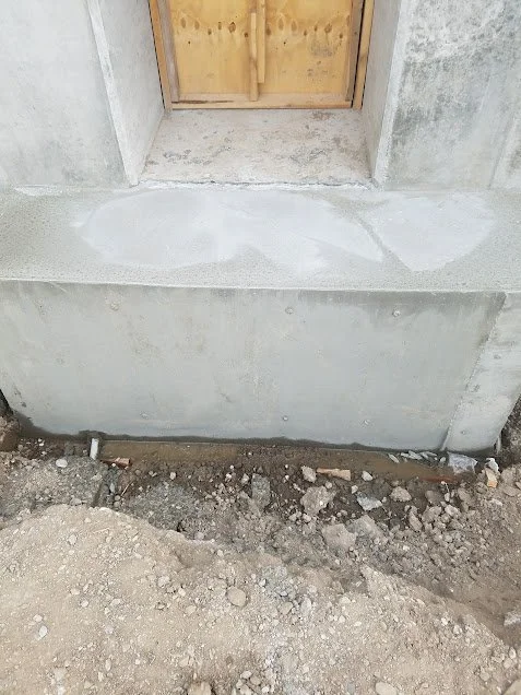 Close-up of a concrete foundation with a door frame, showing unfinished construction with exposed dirt and debris at the base.