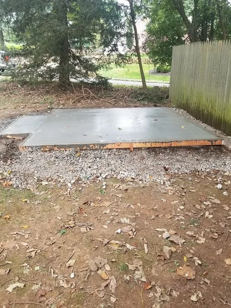 Freshly poured concrete slab on a construction site, surrounded by dirt and gravel, with trees and a wooden fence in the background.