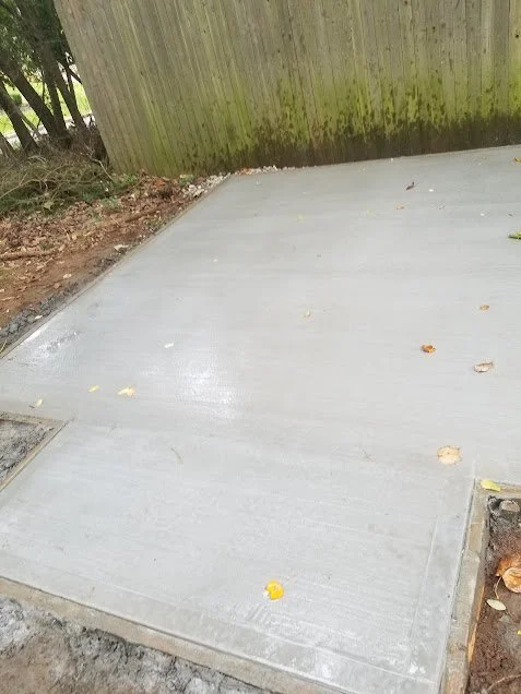 Recently poured concrete patio next to a wooden fence with moss growth at the base, surrounded by dirt and fallen leaves.