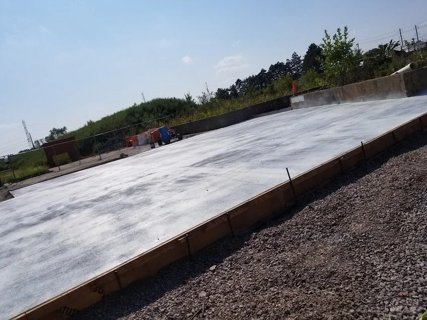 Construction site with a concrete slab, orange and blue barrels, and orange traffic cone, overlooking grassy area with trees and power lines.