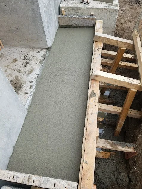 Freshly poured concrete on a construction site, bordered by wooden formwork, with partially completed concrete walls in the background.