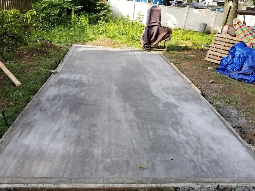 Freshly poured concrete slab in a backyard, surrounded by construction materials and greenery.