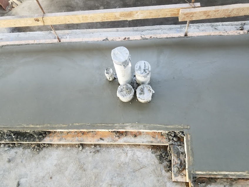 Concrete surface with five protruding metal pipes of varying heights, used as anchors or supports, on a construction site with wooden framing in the background.