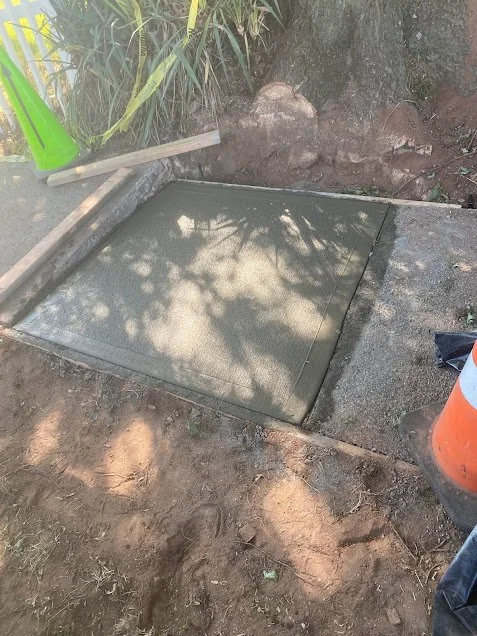 Freshly poured concrete slab surrounded by dirt, with construction tools nearby and foliage casting shade.
