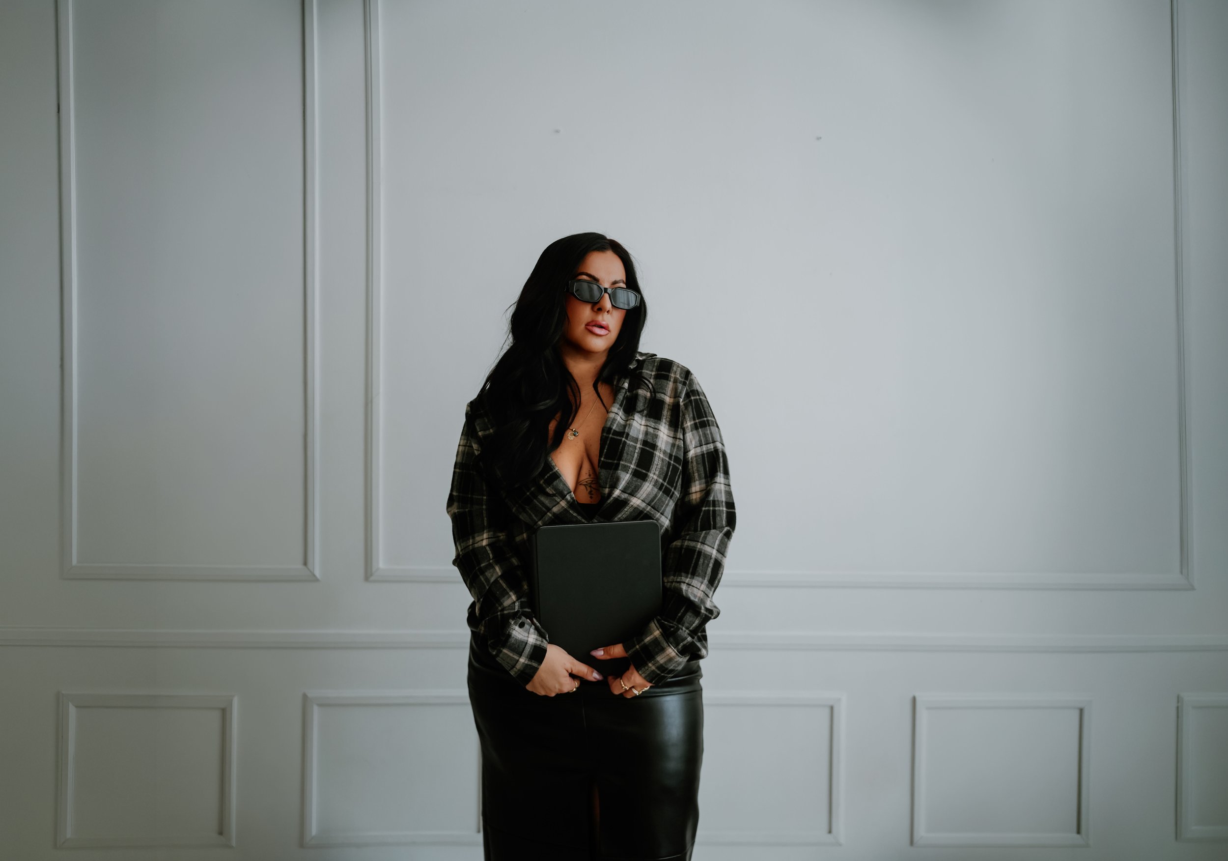 Woman with long black hair, wearing sunglasses, a plaid blazer, and leather skirt, holding a black folder, standing against a white paneled wall.
