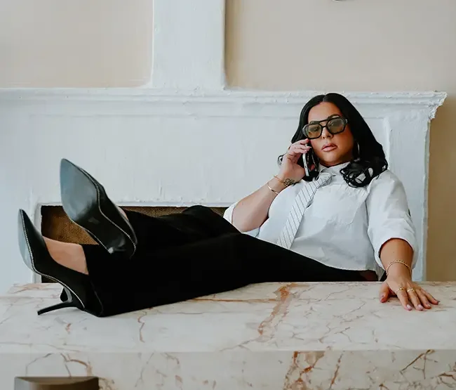 Woman in business attire with dark hair, wearing sunglasses and speaking on cellphone, lounging on a marble-topped table in a bright room.
