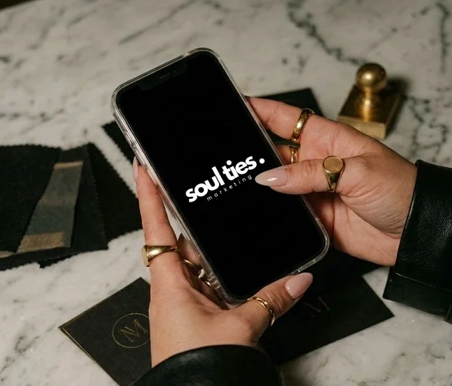 Hands holding a smartphone with the Soul Ties Marketing logo on the screen, standing on a white marble surface with black and gold objects nearby.