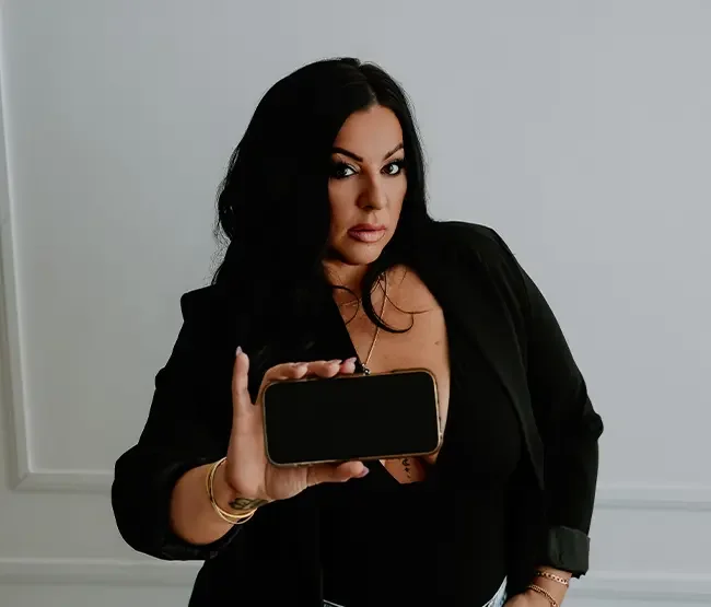 Woman with long dark hair looking at the camera while holding a smartphone in front of her with the screen facing outward, wearing a black blazer and jewelry, against a plain light-gray wall background.