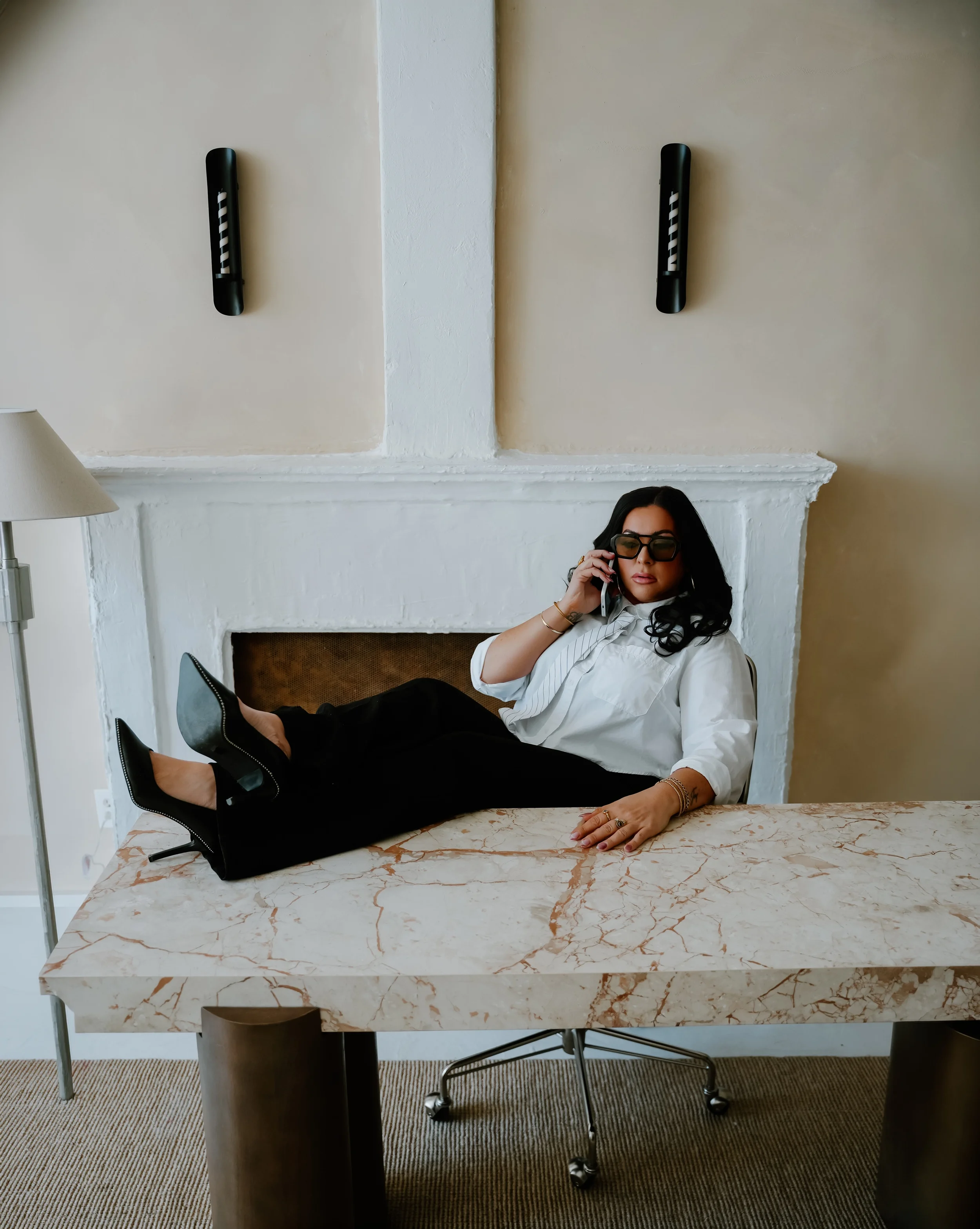 Woman in white blouse and black pants lying on a marble desk, talking on her cellphone, wearing sunglasses, black high heels, in an office or hotel room.