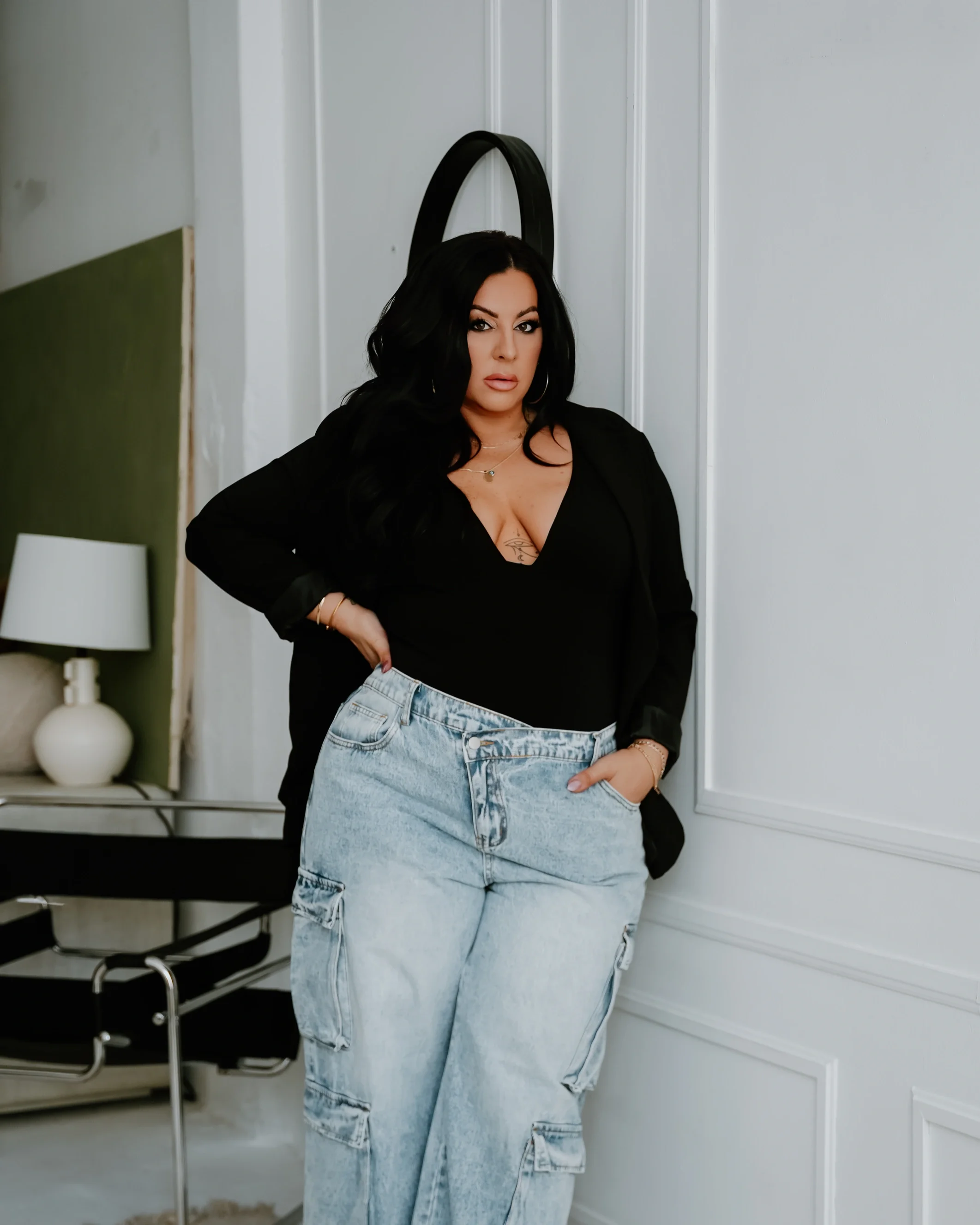 Woman standing in a modern room with white walls, wearing a black top, black blazer, and light blue cargo jeans, with dark hair and makeup.