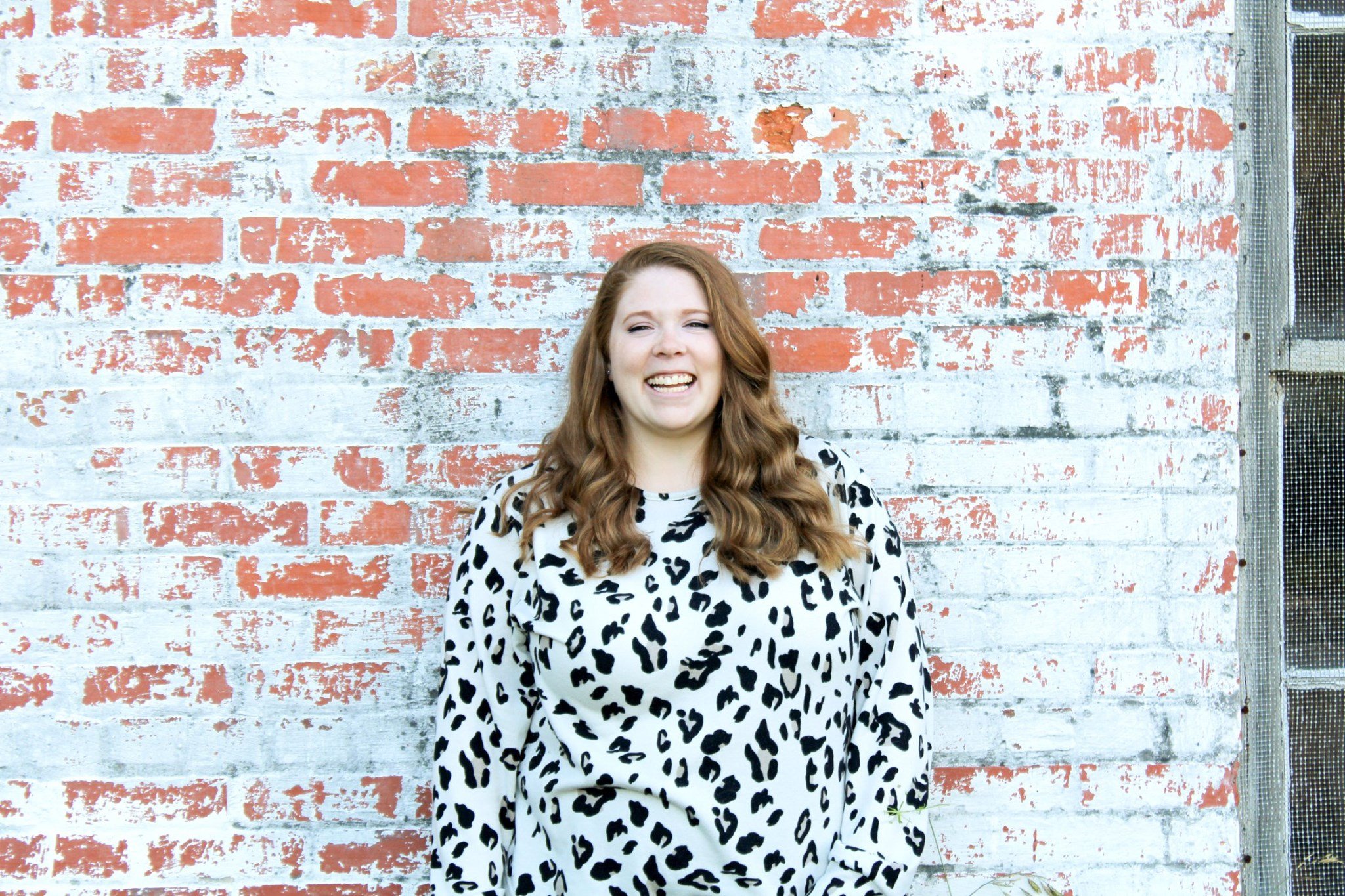 A woman with long curly red hair smiling, standing in front of a weathered brick wall painted white and orange.