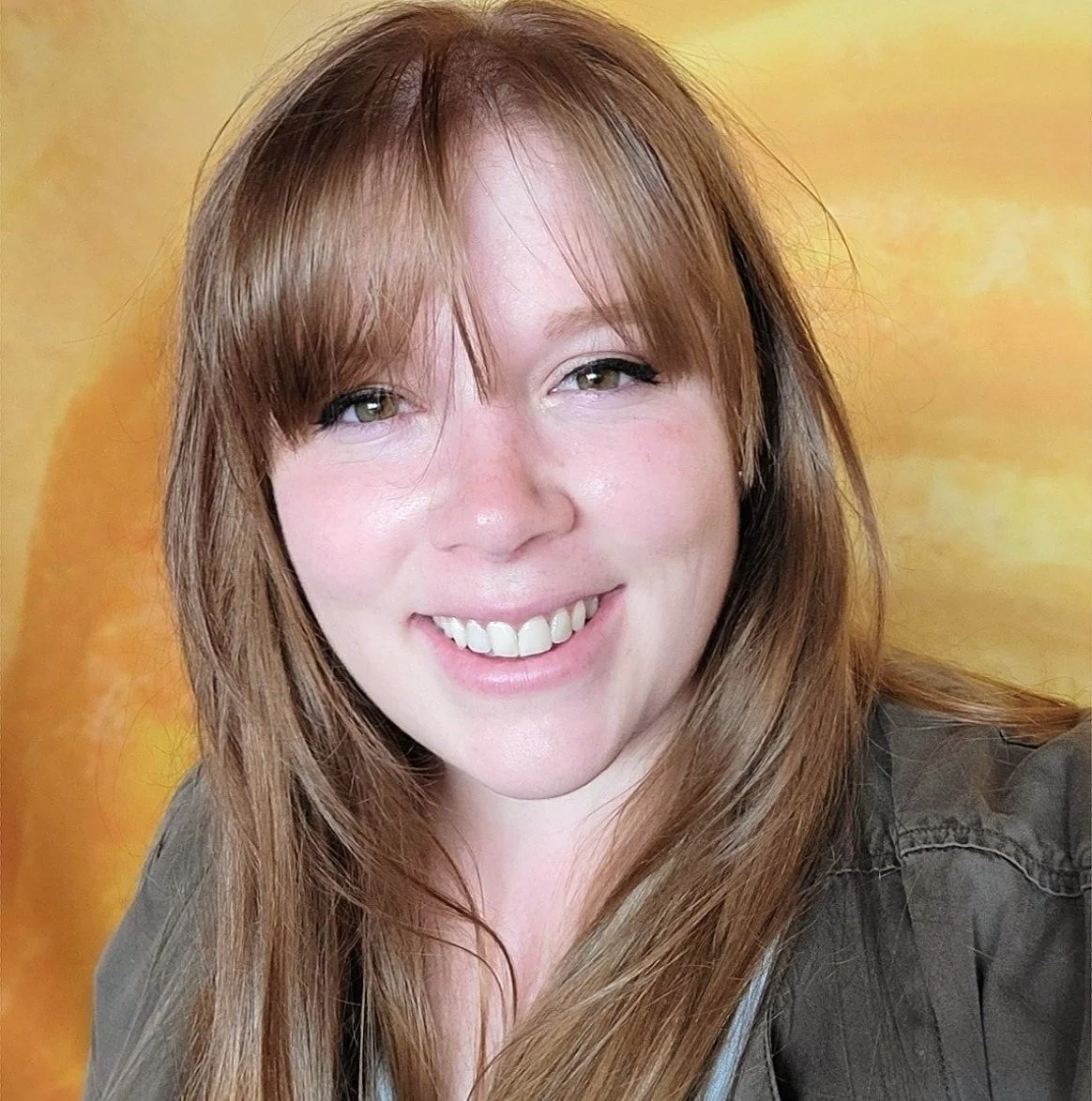 Close-up of a woman with long red hair and bangs, smiling with visible teeth, against a yellow background.