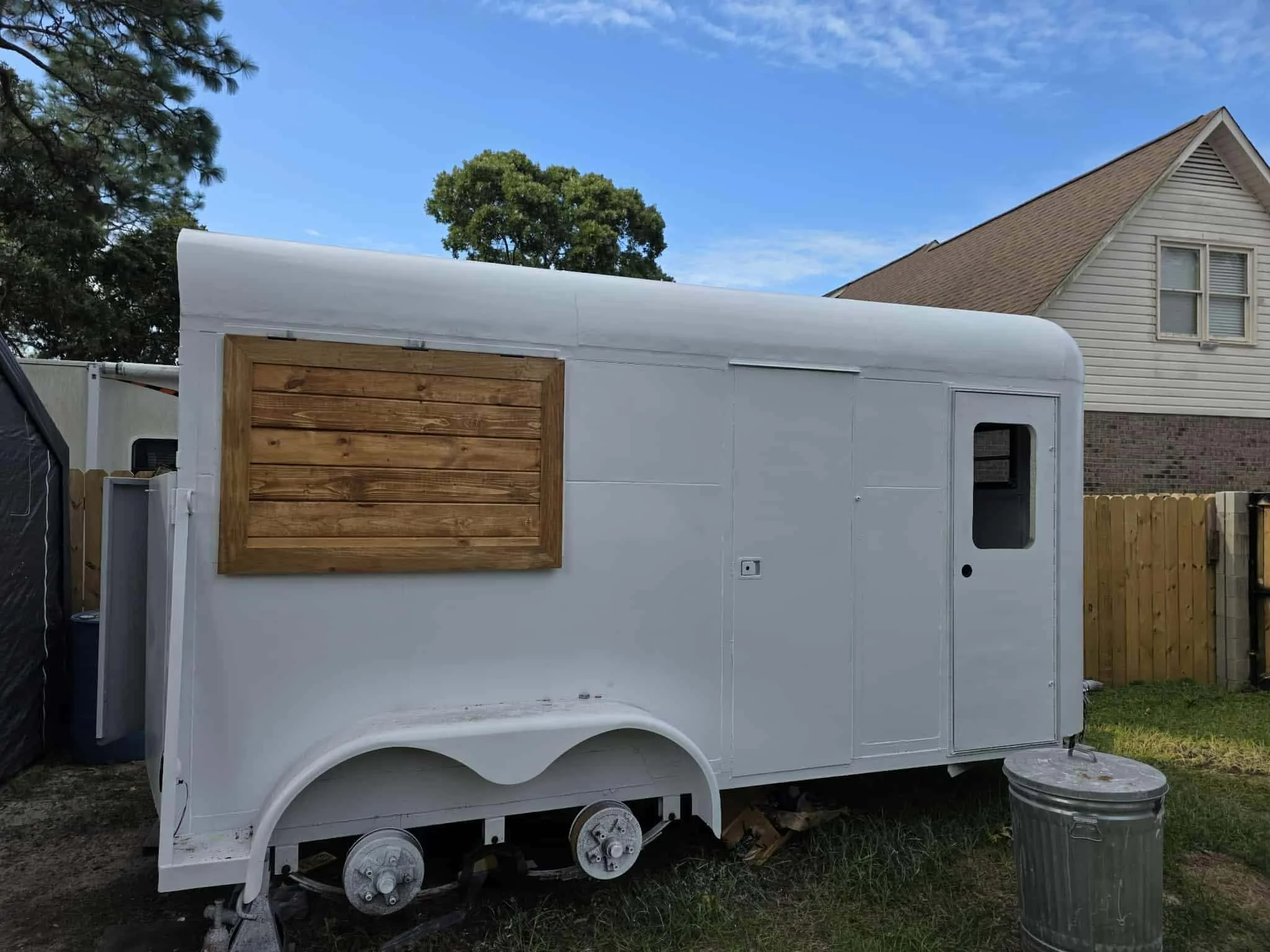 White food truck with a wooden window shutter, parked on grass near a house with beige siding and a wooden fence, under a blue sky with clouds.