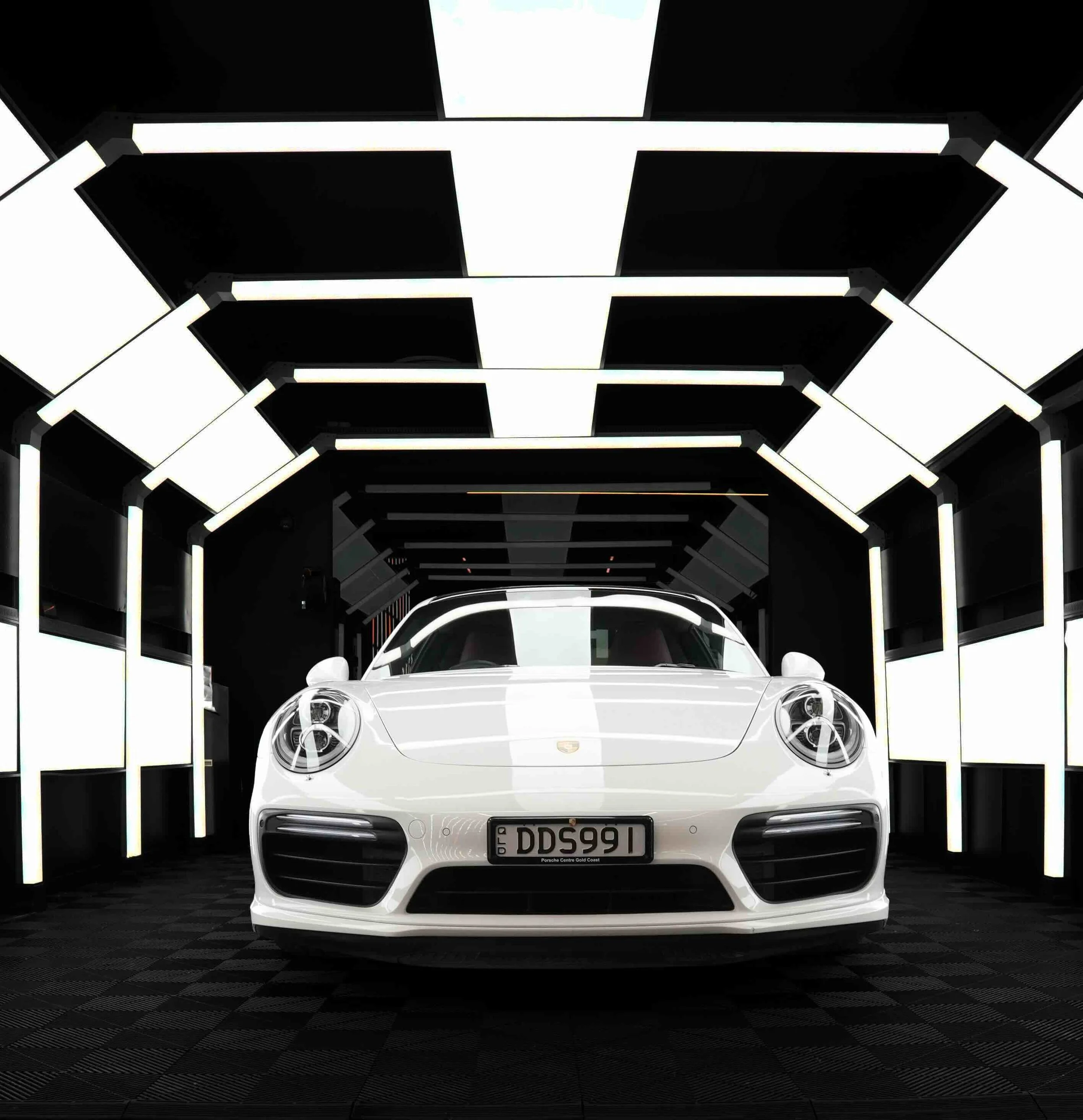 A white Porsche sports car parked in a modern, illuminated garage with black walls and white lighting panels on the ceiling and walls.