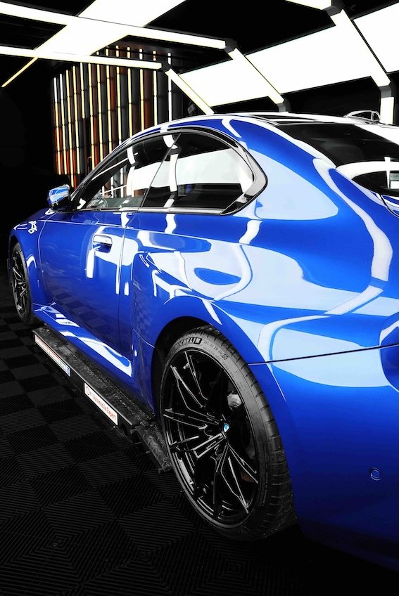A shiny blue sports car displayed in a modern showroom with black and white geometric ceiling panels and wooden accent wall.