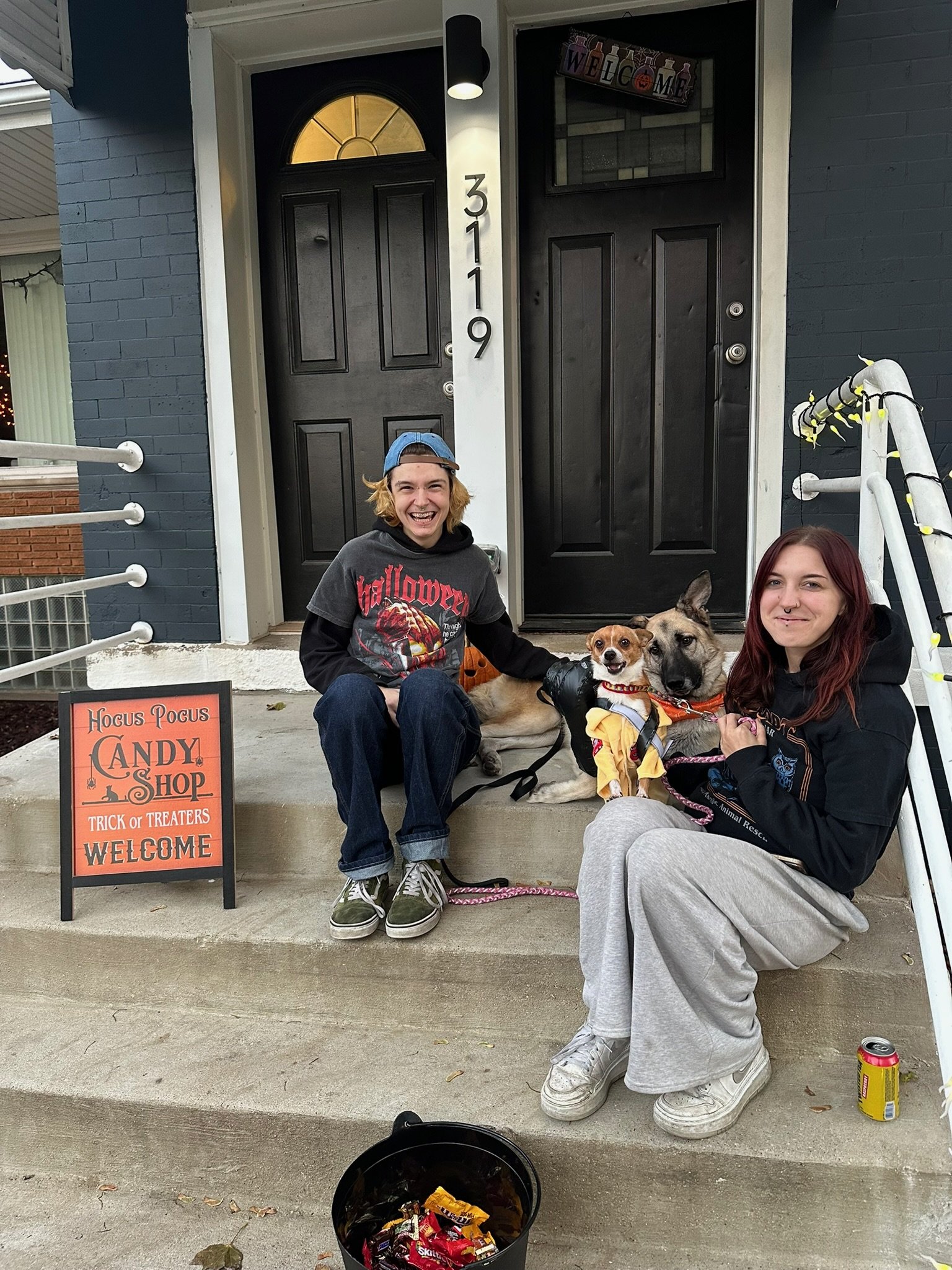 Two people sitting on front steps with two dogs, decorated for Halloween, with a sign for a candy shop and Halloween treats, and a black bucket of candy in front.