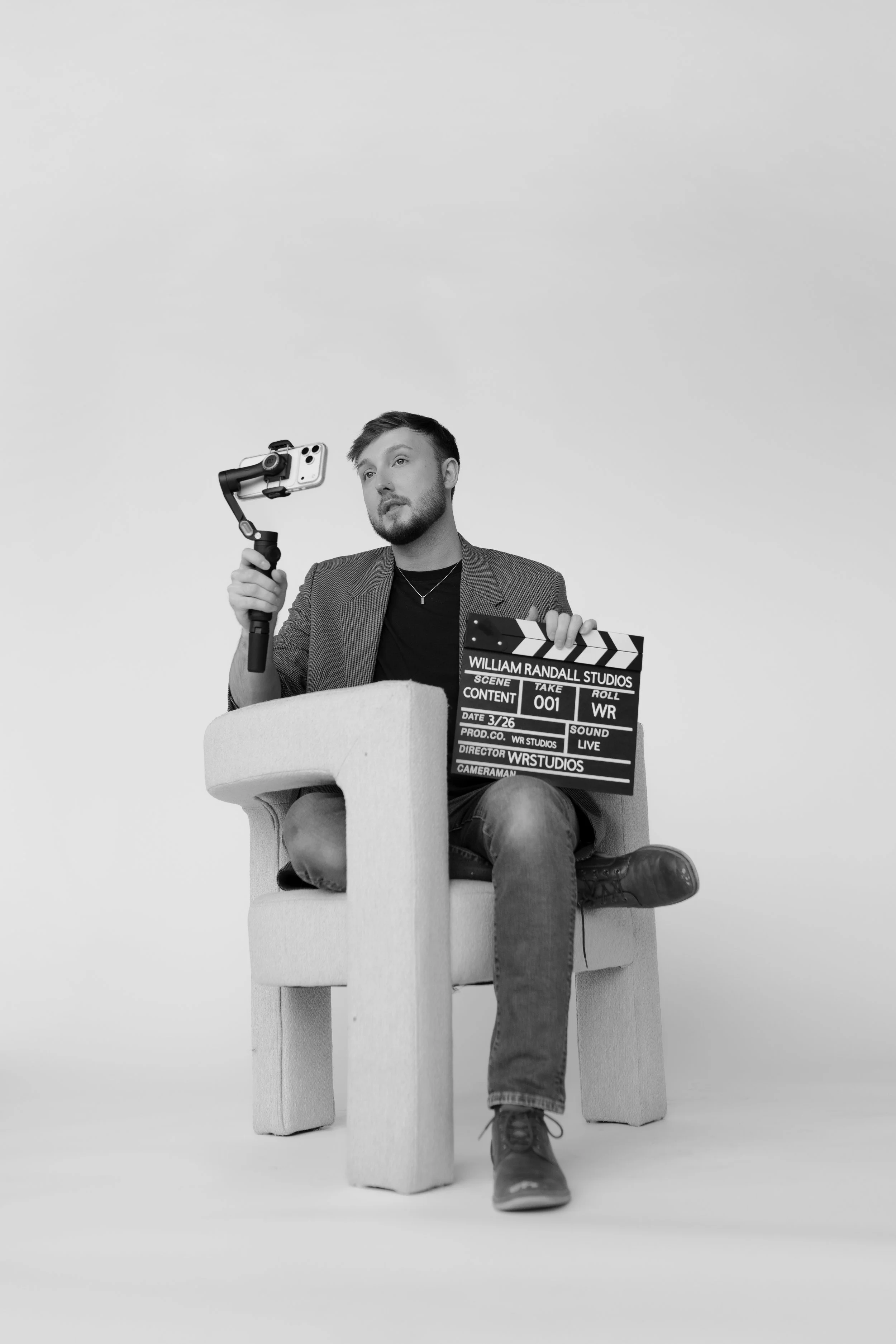 William Randall holding a camera setup and clapperboard while seated during a content shoot in black and white.