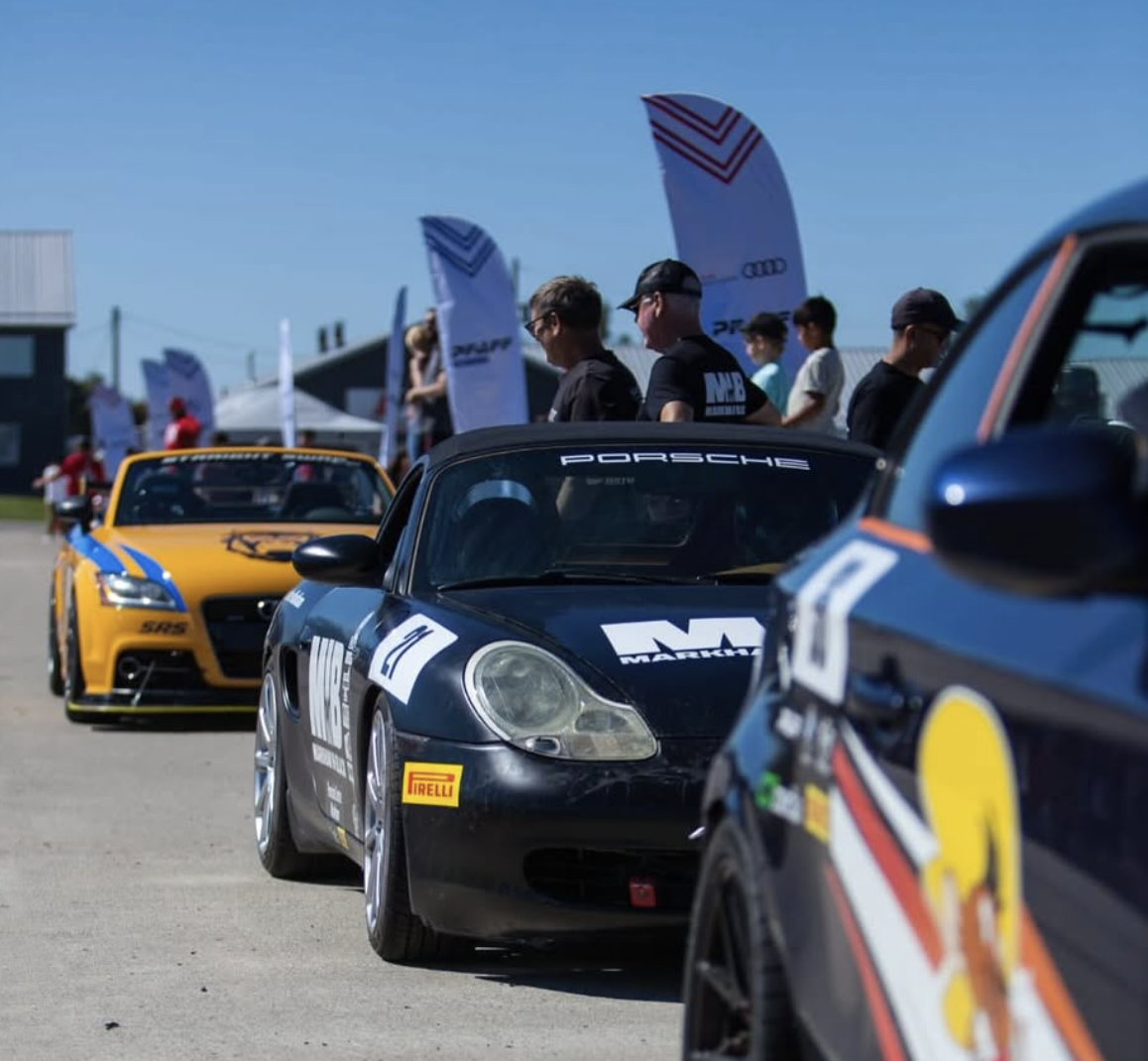 Lineup of racing cars at a motorsport event, with people standing and flags in the background.