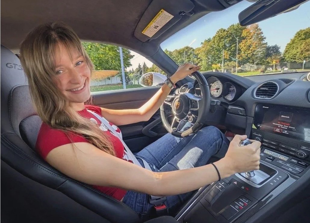 Amanda sitting in the driver's seat of a sports car, holding the gear shift with her right hand, with a view of trees and a street outside through the window.