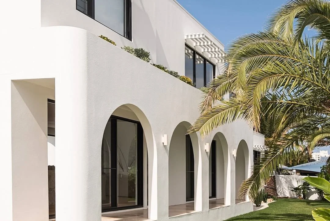 Modern white house with arched doorways, large windows, and a rooftop garden, with palm trees and a well-maintained lawn.