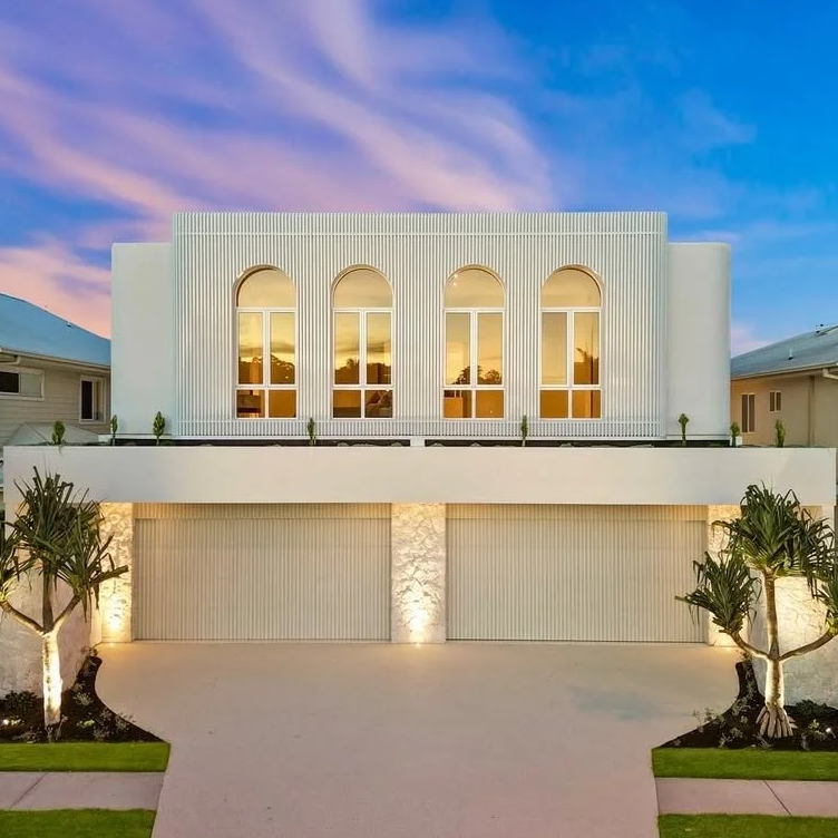 Modern white house with three tall arched windows on the upper floor, a driveway in front, and landscaped trees and grass on either side, during sunset.