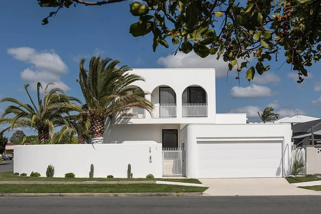 A modern white two-story house with arched windows, a white fence, and palm trees in a suburban neighborhood under a blue sky with scattered clouds.