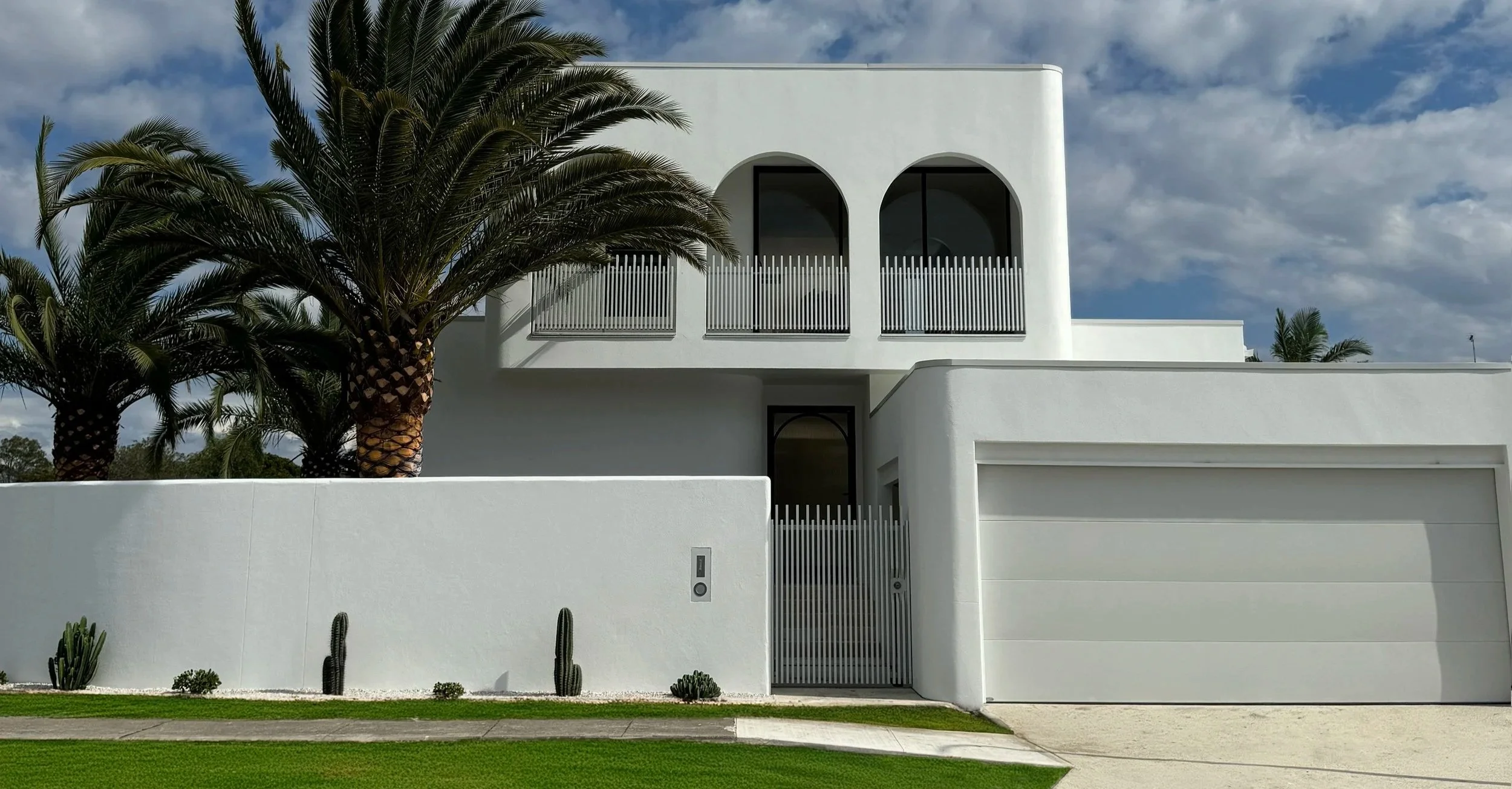 Modern white two-story house with arched windows, black accents, a large garage door, surrounded by palm trees and desert plants, with a grass lawn and a sidewalk in the front.