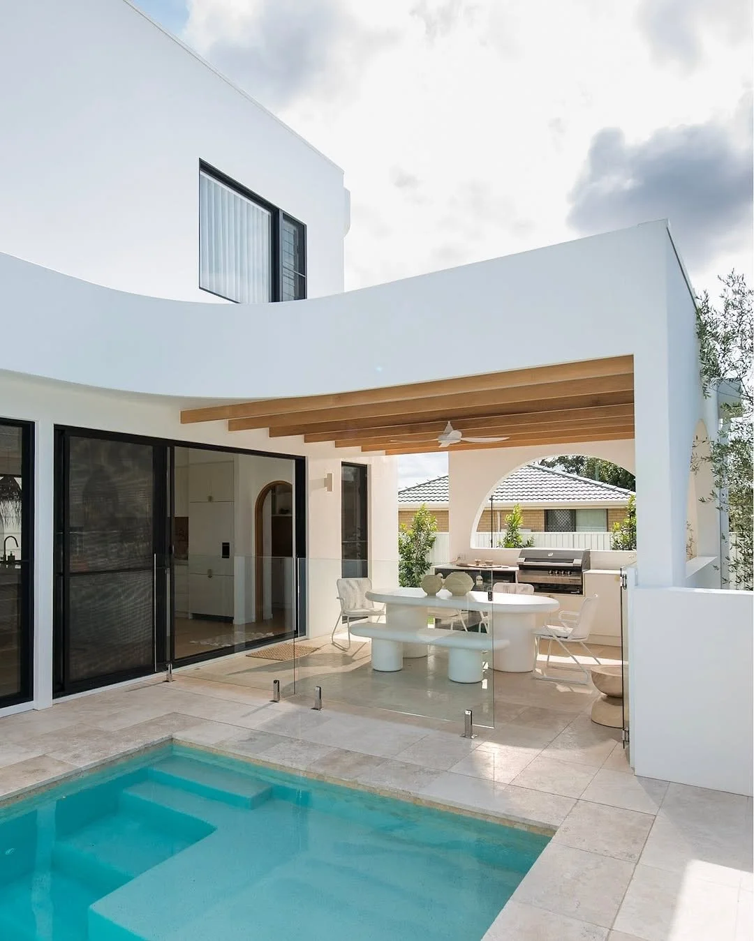 Modern white house patio with a swimming pool, outdoor dining area, and built-in grill, featuring glass fencing, wooden beams, and minimalistic decor.