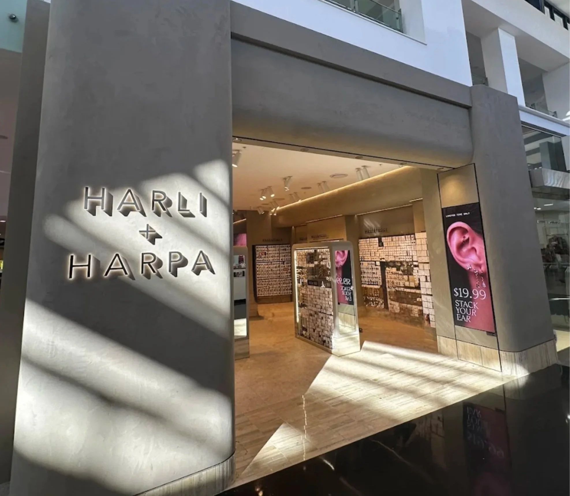 An entrance to a Harli + Harpa store with illuminated sign on concrete wall and digital advertising display on side. Inside, store walls display various items and advertisements.