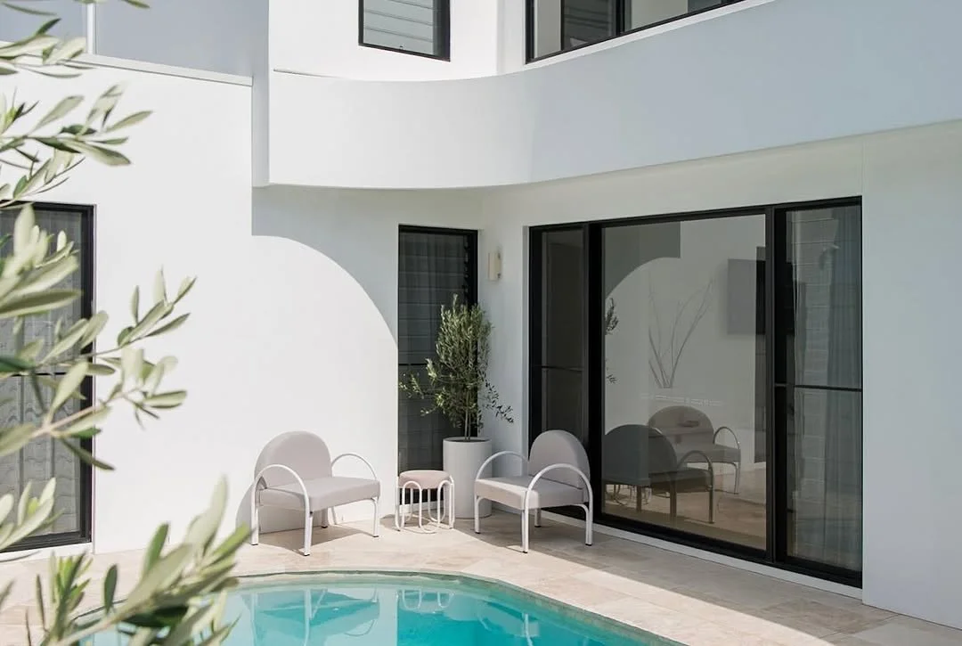 Modern residential courtyard with a swimming pool, white patio furniture, potted plants, and sliding glass doors, with a white building backdrop.