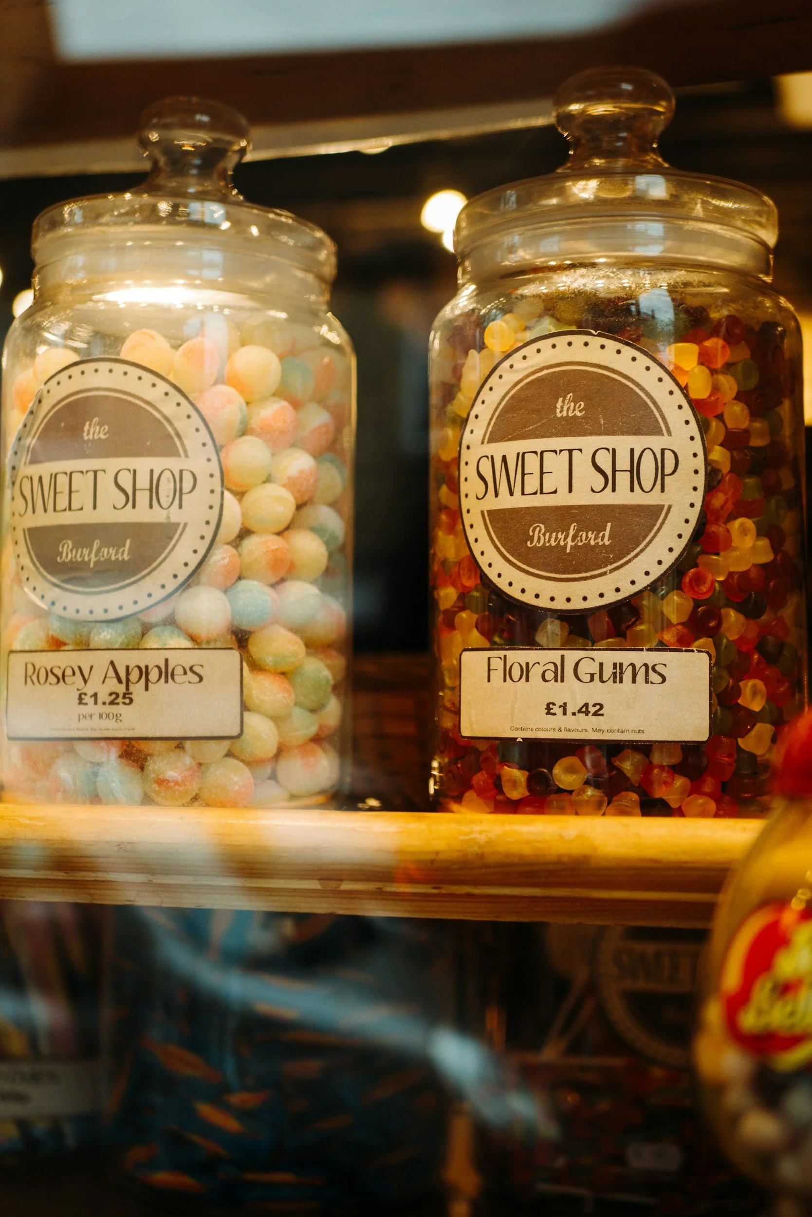 Two glass jars labeled 'the Sweet Shop Burland' on a wooden shelf. The left jar contains pastel-colored round candies titled 'Rosey Apples,' and the right jar contains multicolored gummy candies named 'Floral Gums.'