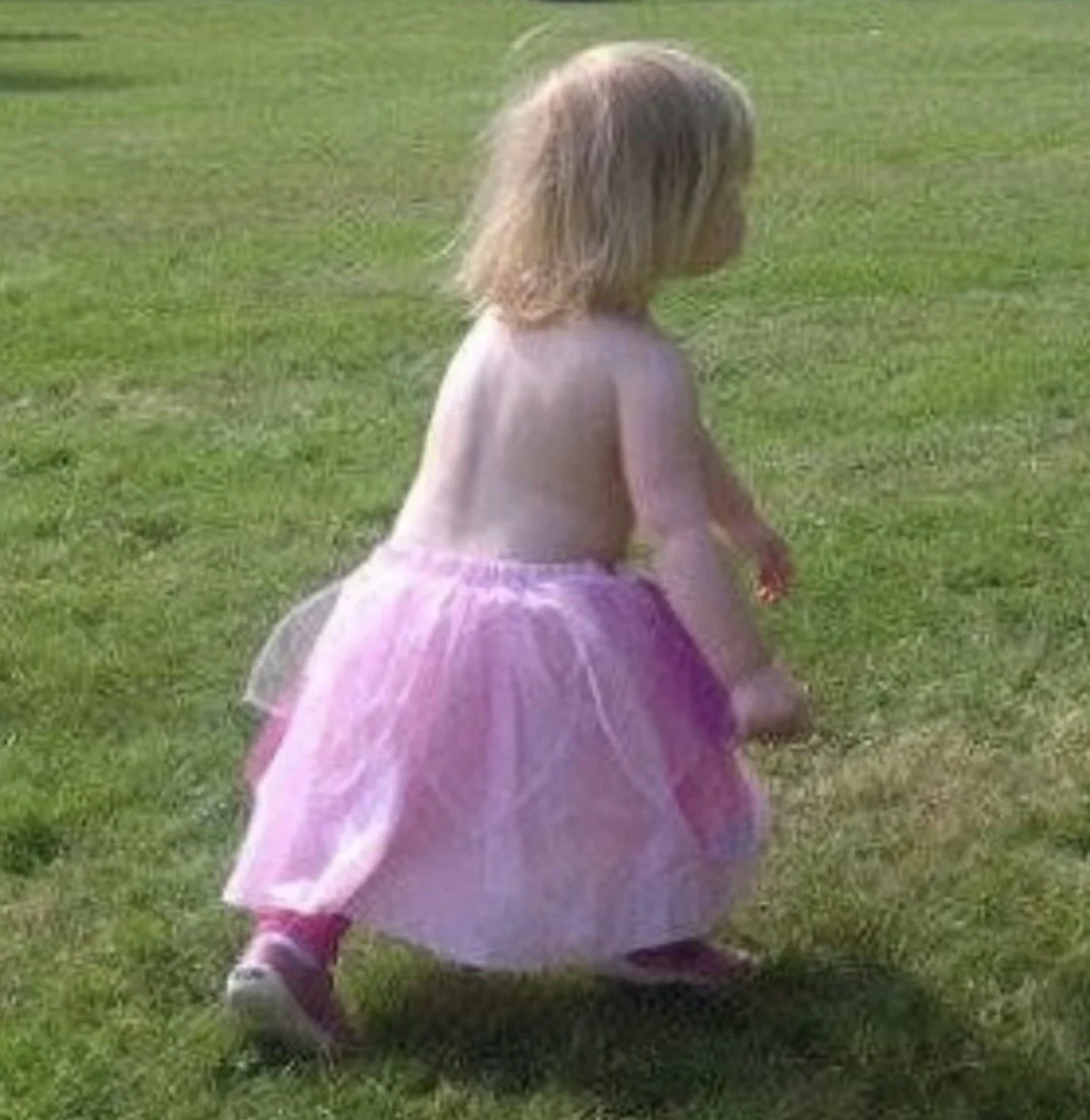 A young girl with blonde hair wearing a pink tutu and t-shirt is marching across the grass with purpose in park.