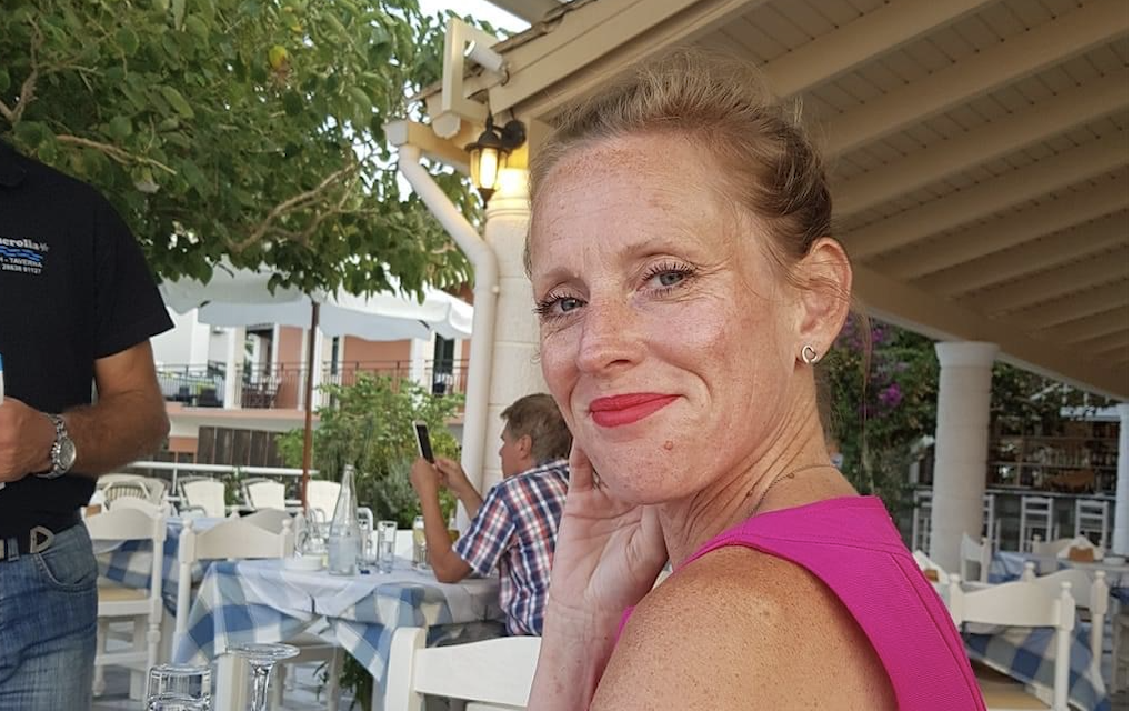 A woman with red hair and freckles smiling at the camera, wearing a pink sleeveless top, sitting at an outdoor restaurant table with a blue and white checkered tablecloth, with other people and a house with a porch in the background.