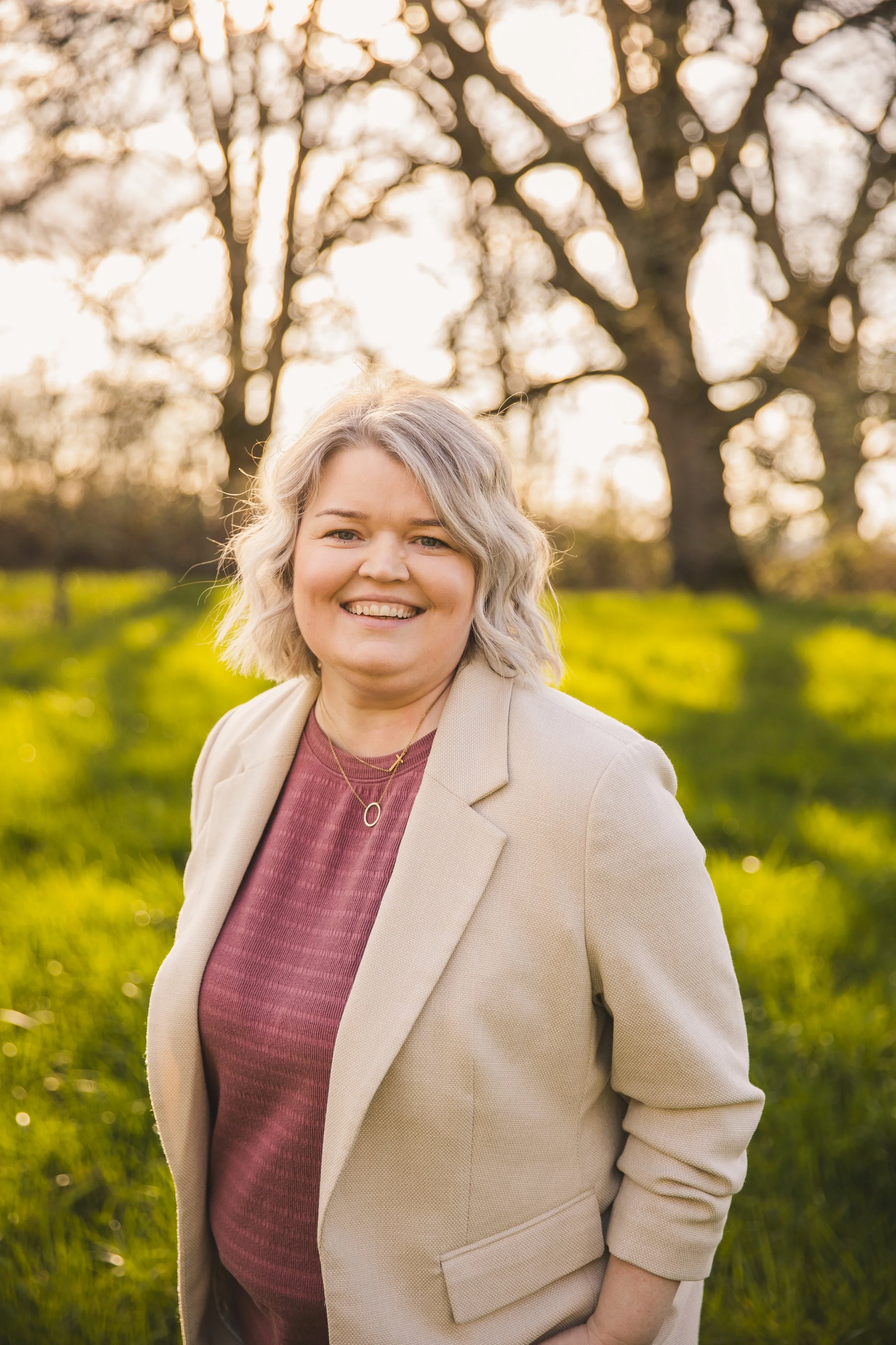 A woman with blonde, curly hair smiling outdoors during sunset, wearing a beige blazer over a maroon top, with a background of trees and green grass.