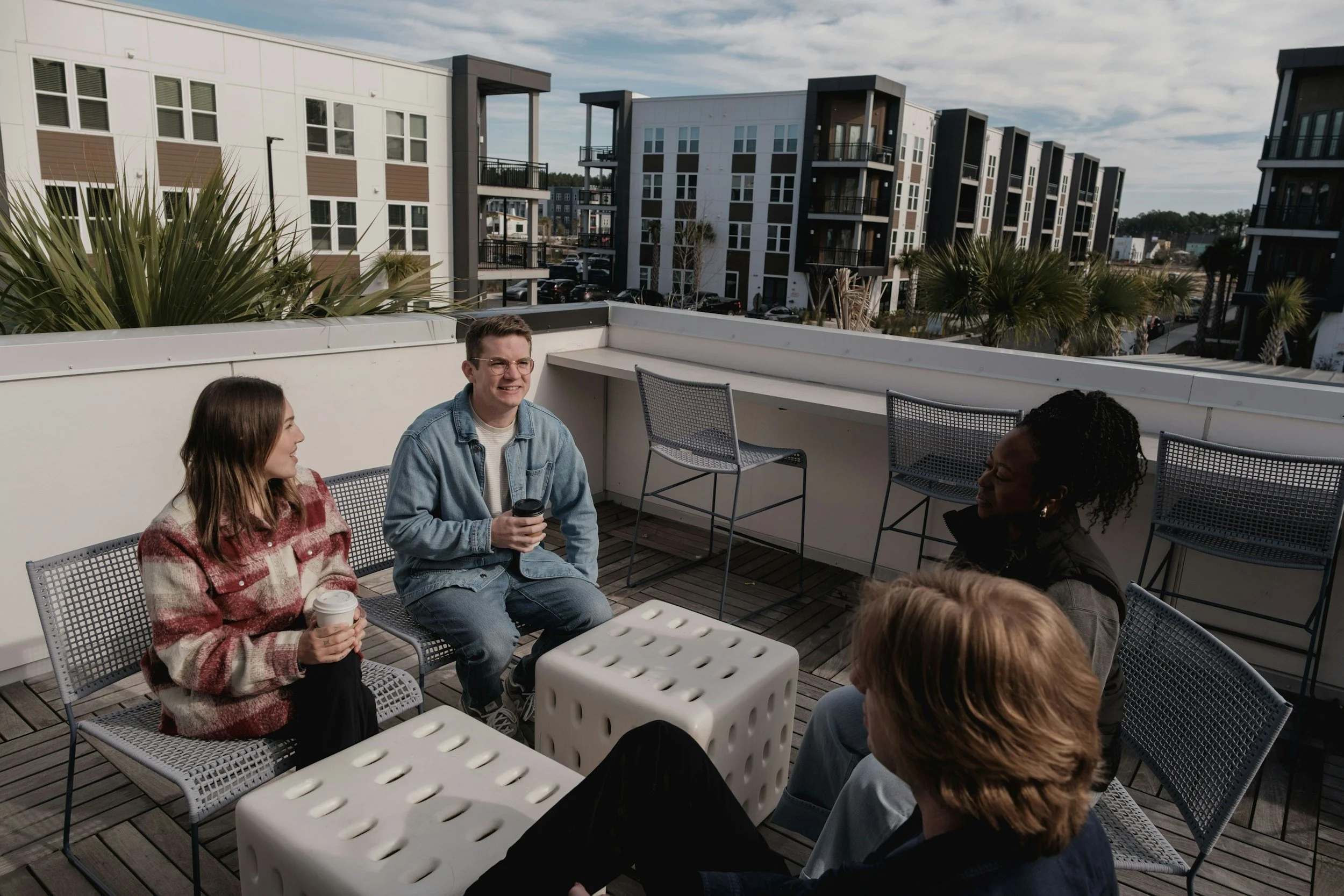 Five friends talking and drinking coffee on a rooftop patio with modern apartment buildings in the background.