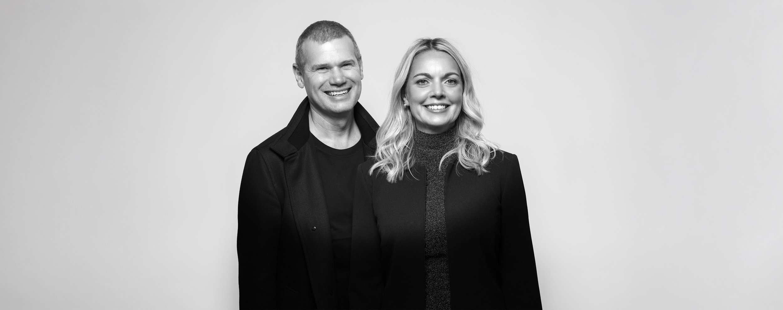 A black and white photograph of a smiling man and woman standing side by side against a plain background, dressed in dark clothing.