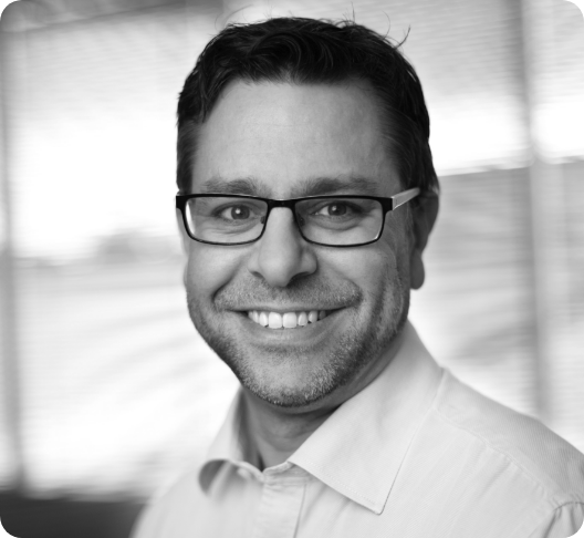 Headshot of a smiling man wearing glasses and a white collared shirt, in a professional setting with blurred background.
