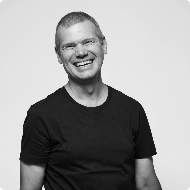 A smiling man with short hair wearing a black t-shirt, standing against a plain light background.